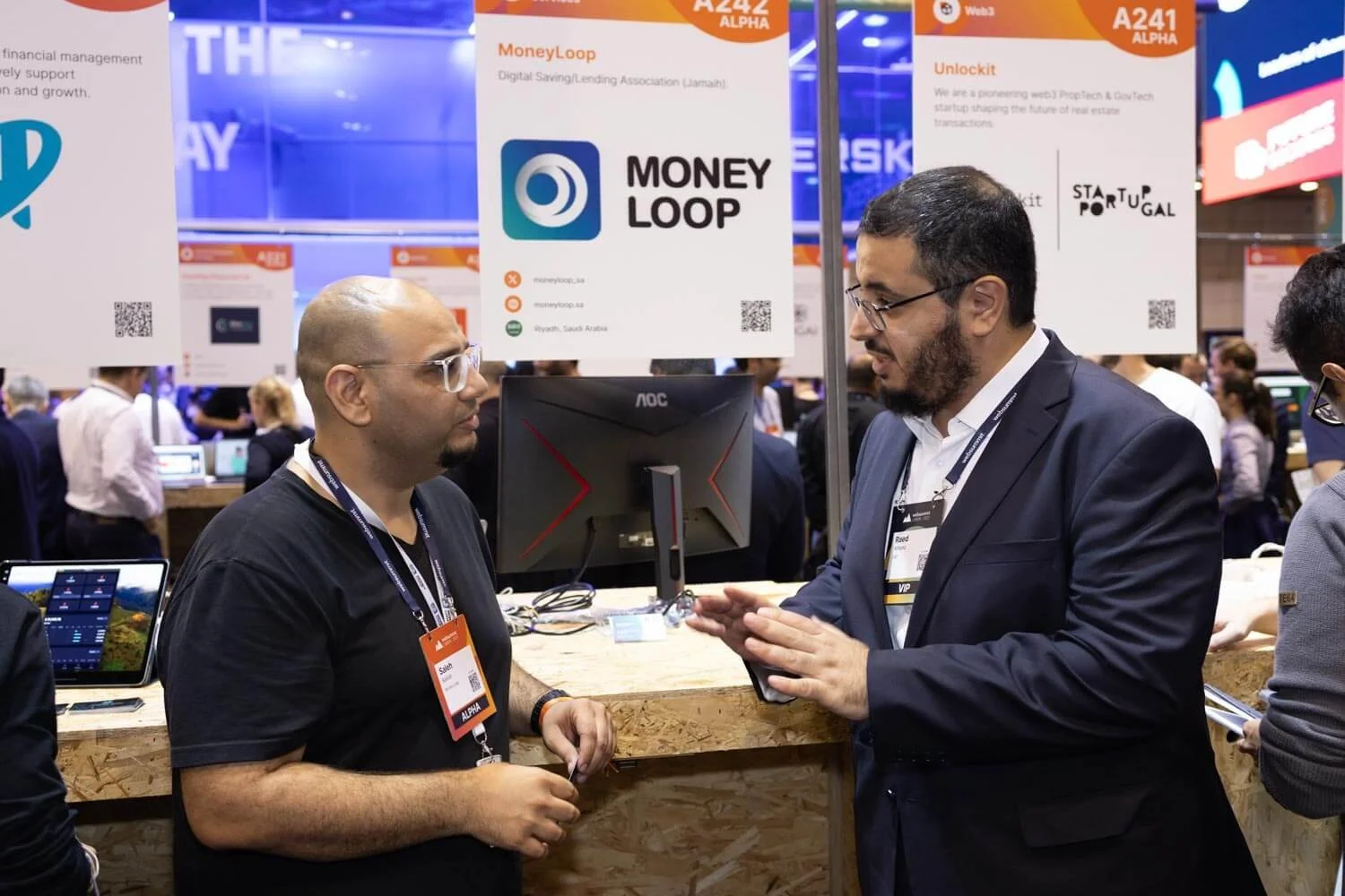 Two men are having a conversation at an exhibition booth with several signs and posters displaying the words 'Money Loop,' 'Unlockit,' and 'StartUp Portugal.' There are monitors and informational materials on a wooden counter in front of them, with a