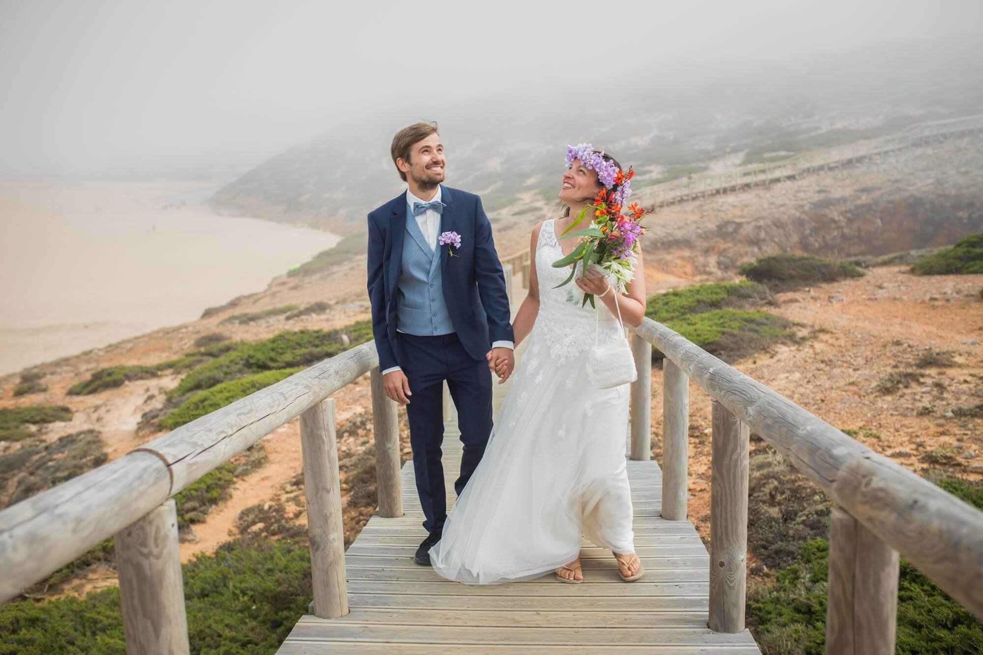 A newlywed couple holding hands on a wooden walkway near the coast, with a foggy beach and cliffs in the background. The bride is in a white dress with a floral crown and bouquet, and the groom is in a blue suit with a bow tie.