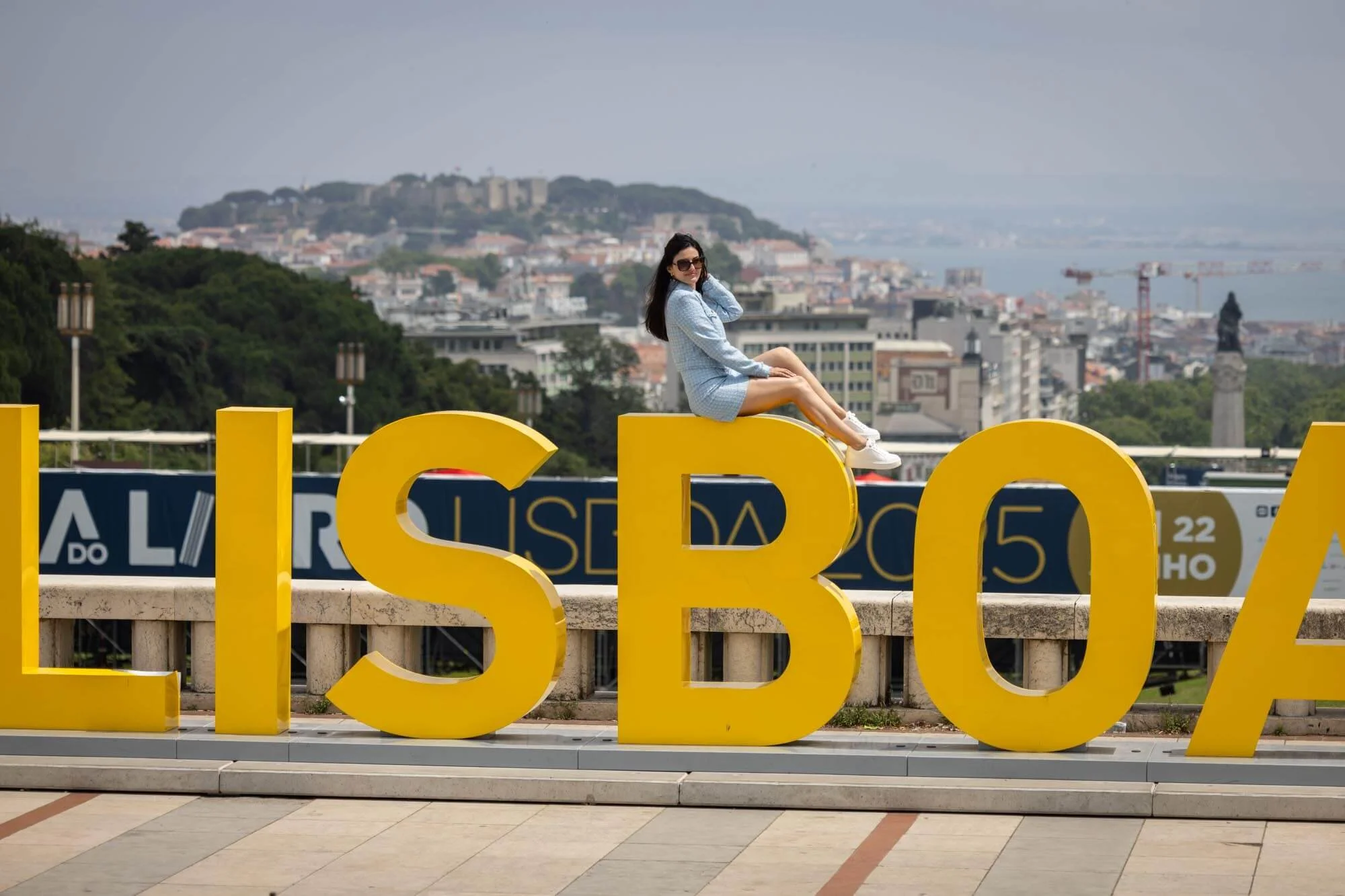 Woman sitting on large yellow letters spelling 'LISBOA' in a public square with cityscape, forested hills, and water in the background.