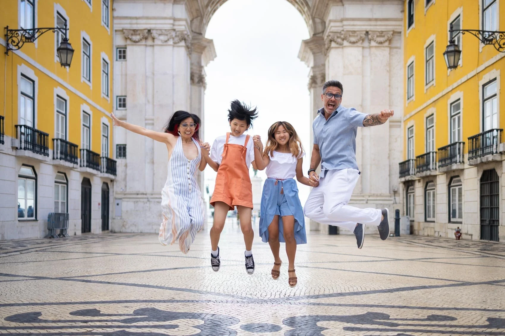 Four people jumping in the air under the Arco de Rua Augusta in Lisbon,  with colorful buildings and archways, smiling and holding hands.