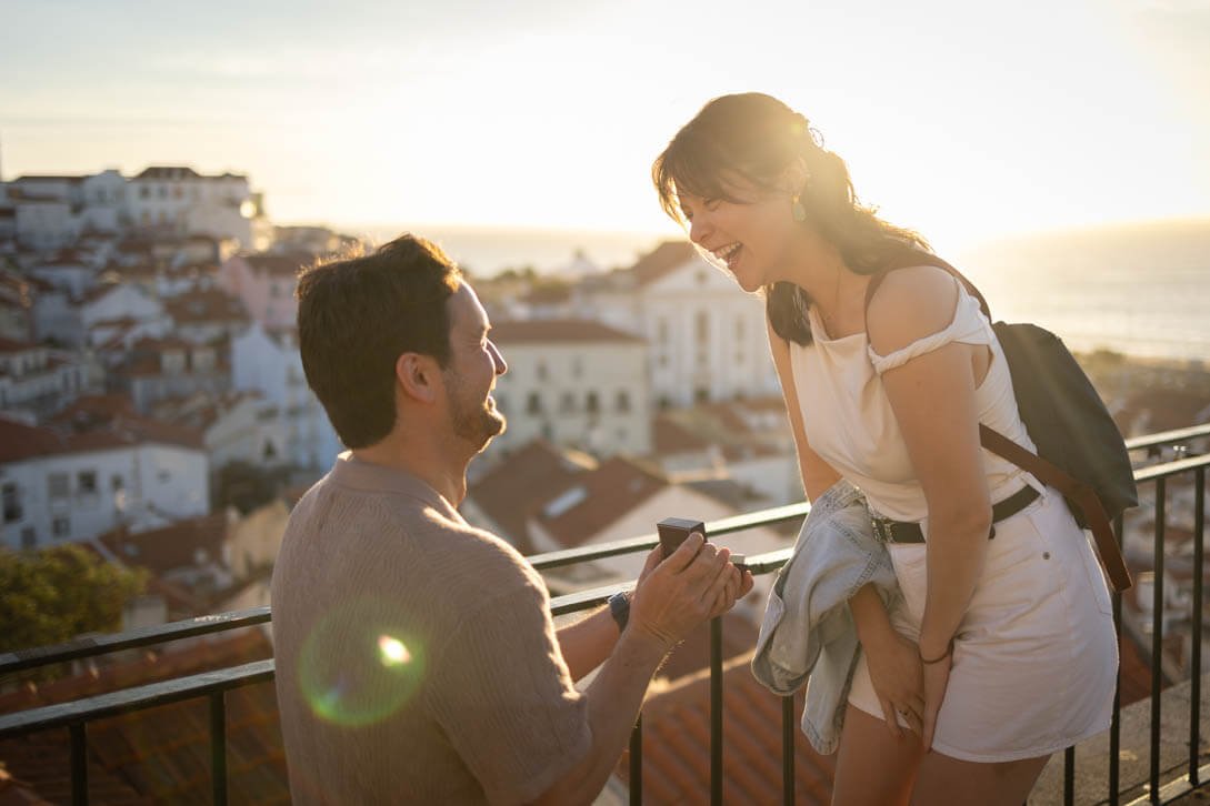 A man proposing to a woman on a balcony at sunset, with a cityscape and ocean in the background, both smiling and laughing.