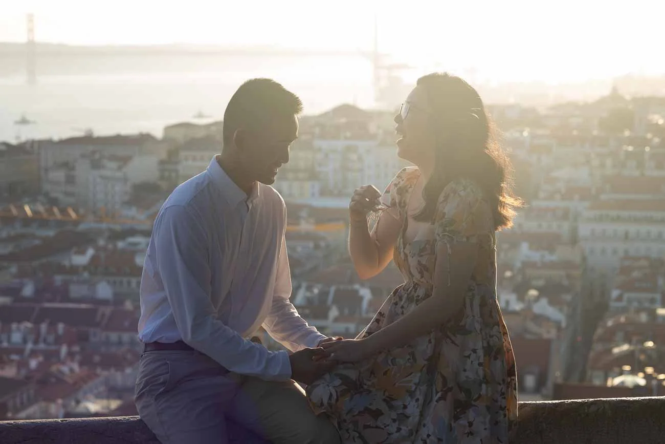 Couple sitting and smiling, with a Lisbon in the the background. Sunset Secret Proposal Photoshoot at São Jorge Castle with a lovely couple from USA.
