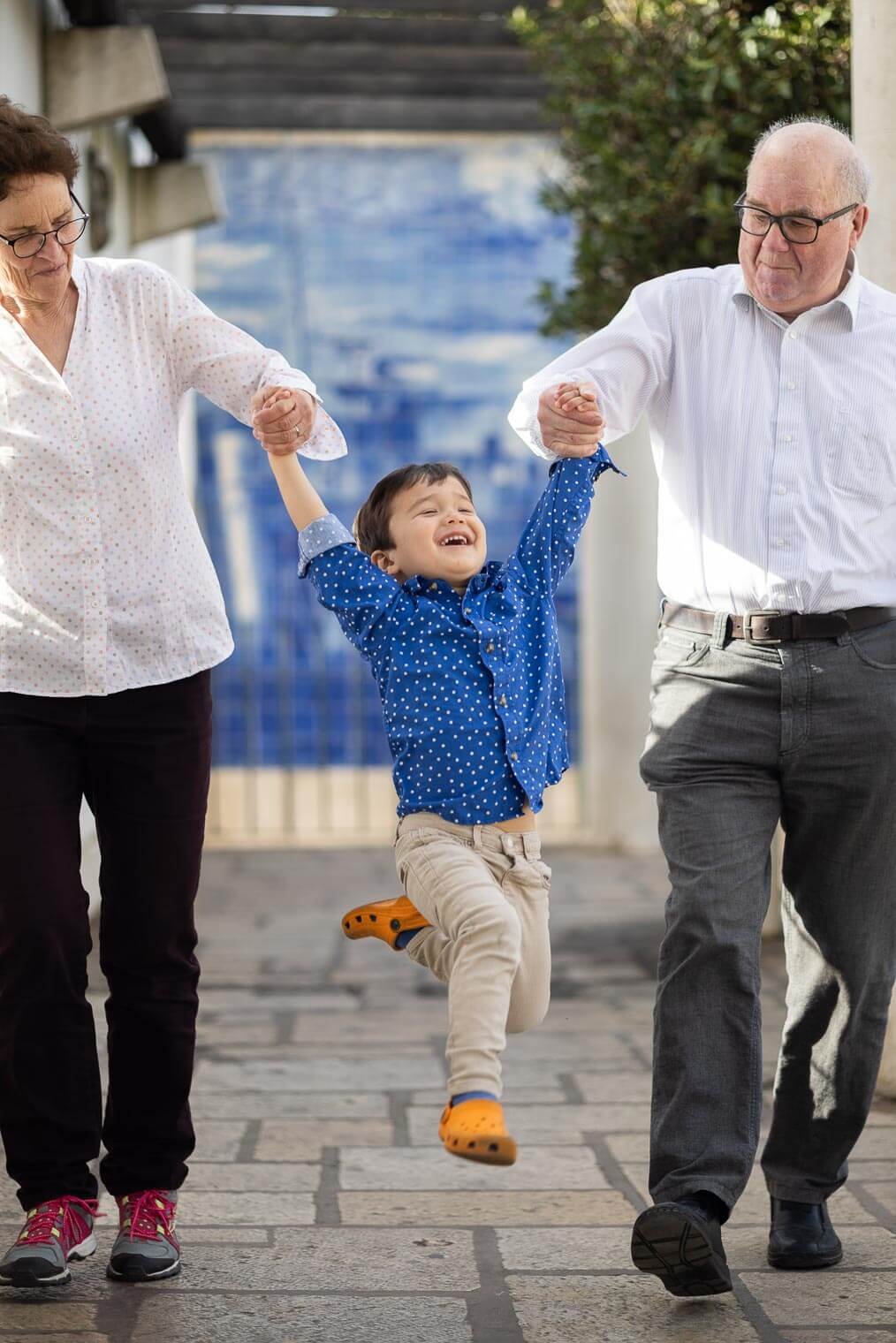 A young boy in a blue polka dot shirt is happily jumping while holding the hands of two older adults, likely his grandparents, as they walk outdoors on a stone pathway.