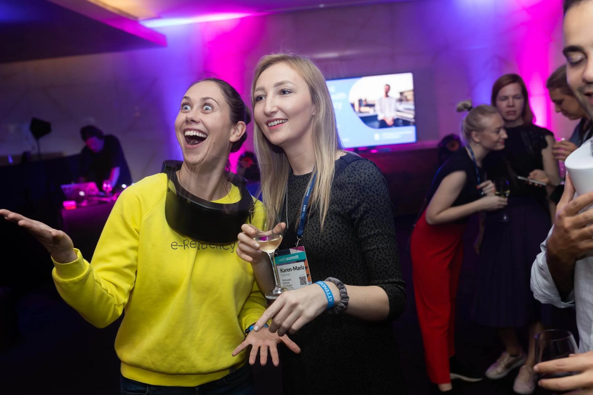 Two women smiling and talking at an event, with one wearing a yellow sweatshirt and the other in black, holding a glass of wine. Other people are in the background, and there are pink and purple lighting effects.