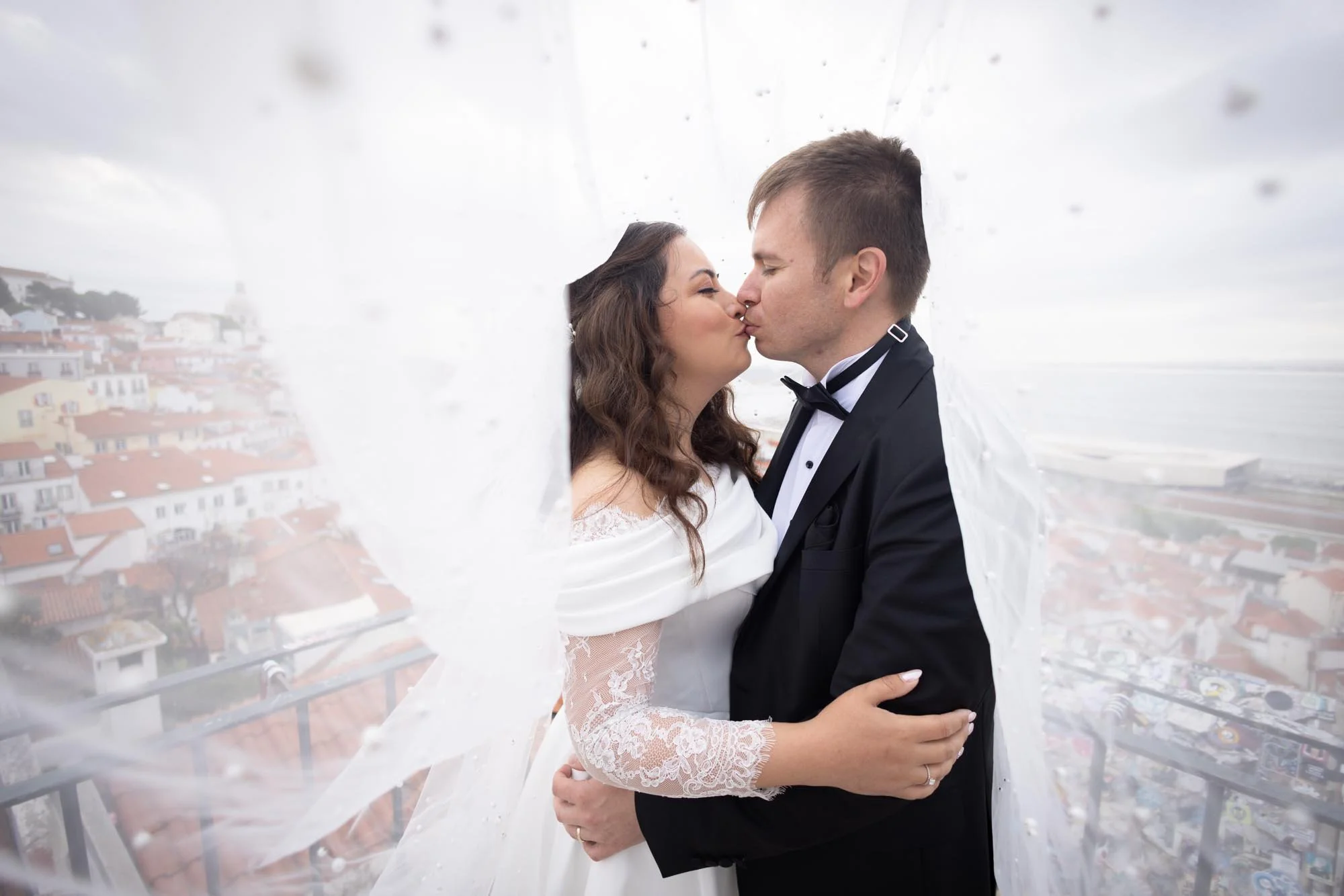 Bride and groom sharing a kiss on a rooftop with city views, the bride in a white lace dress and the groom in a tuxedo.