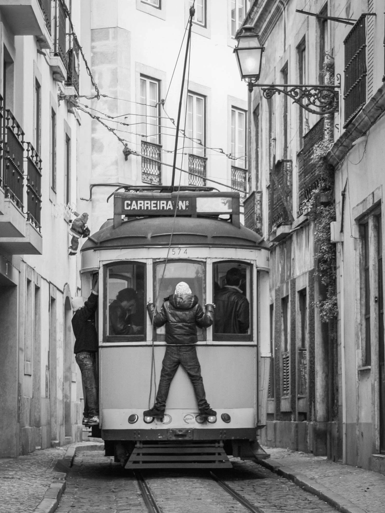 Black and white photo of an old tram with three kids hanging onto the front, on a narrow street with apartment buildings and balconies.