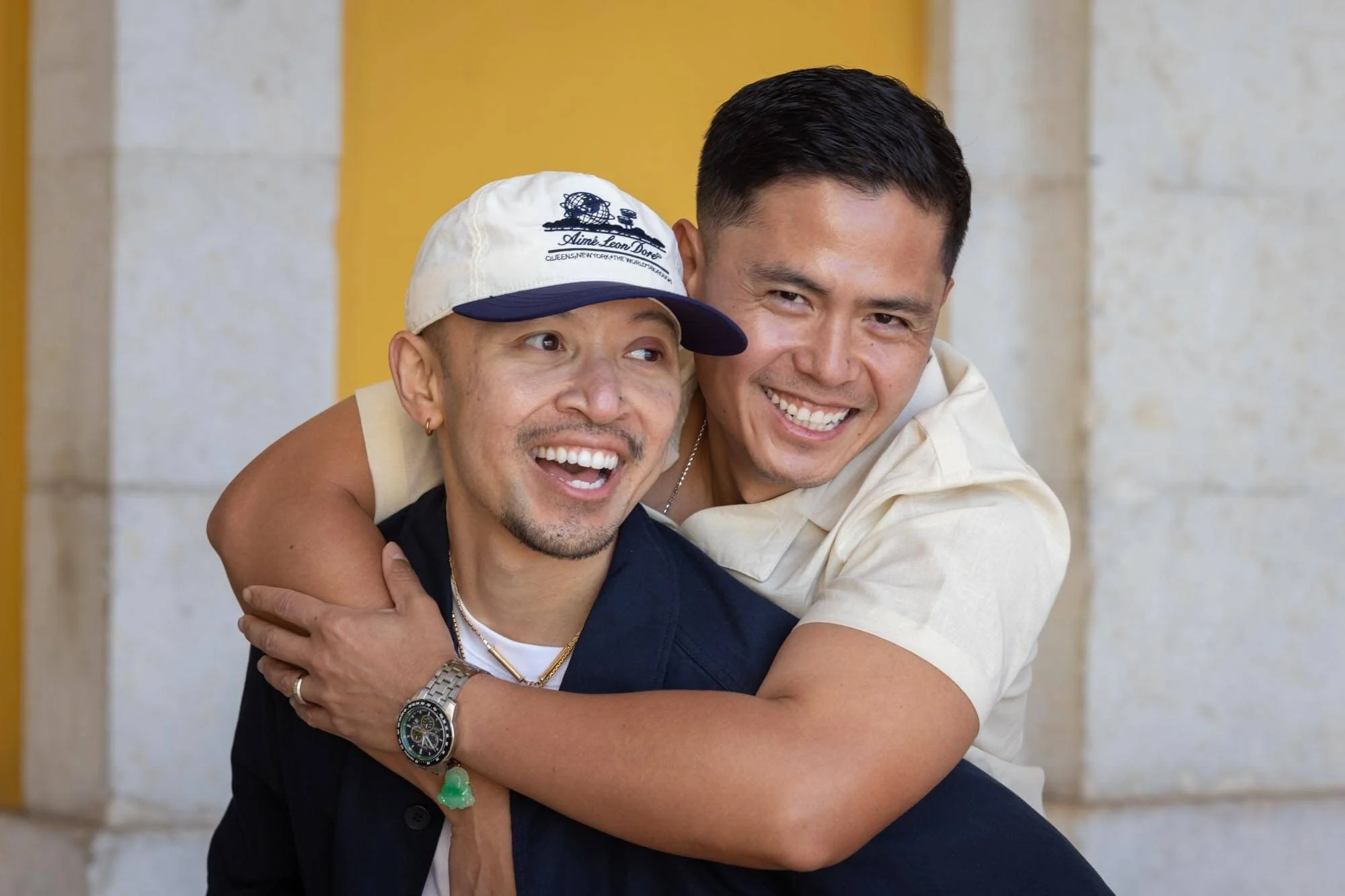 Two men smiling and hugging each other, one wearing a white cap and the other with short dark hair, both dressed casually, standing against a beige and yellow background.