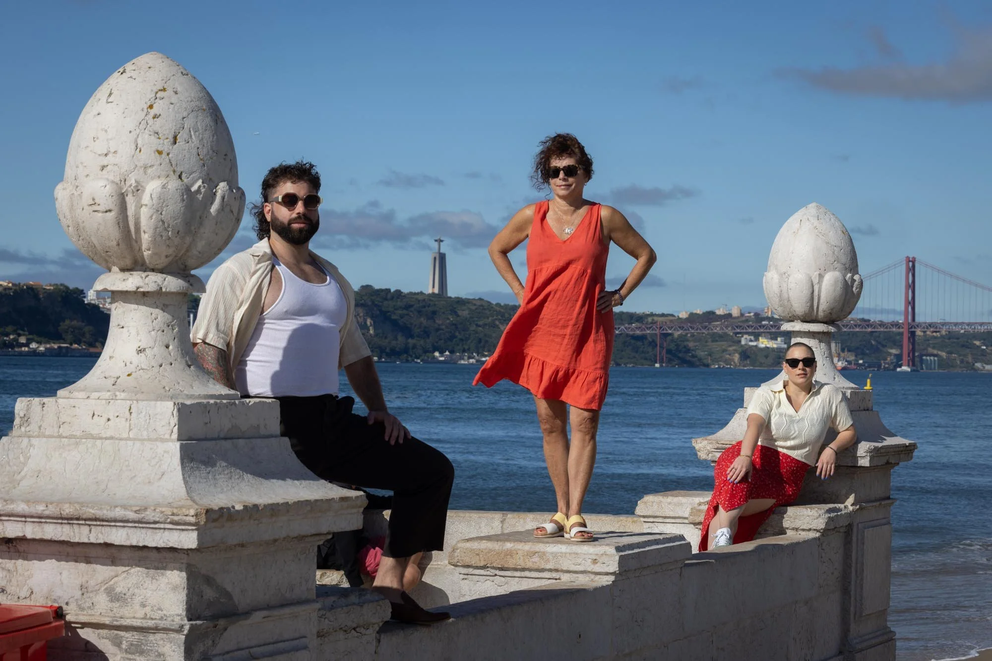 Three people posing on a stone structure by the water, with a bridge and the Christ the Redeemer statue in the background.
