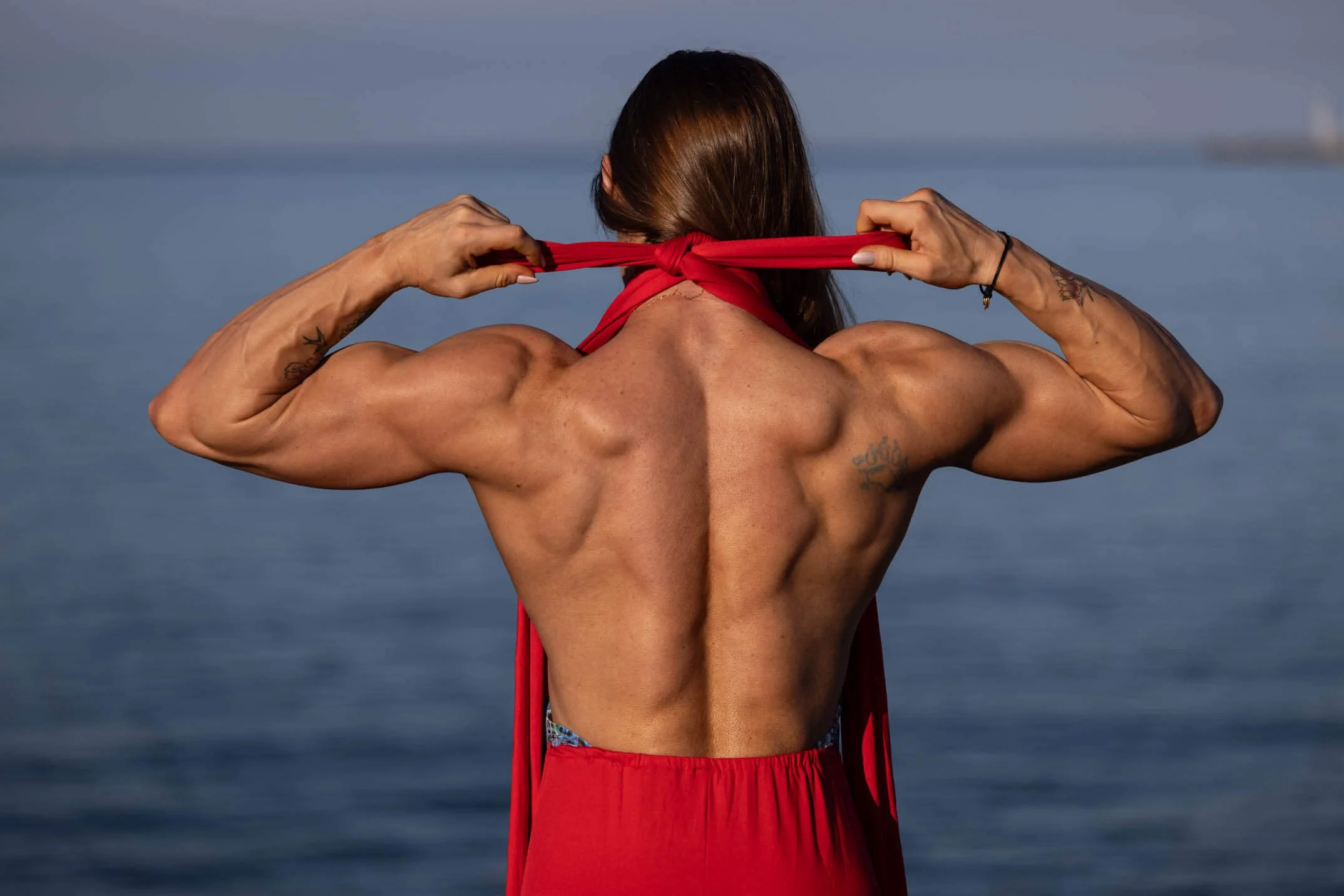 Back view of a muscular woman tying a red scarf around her neck near a body of water.