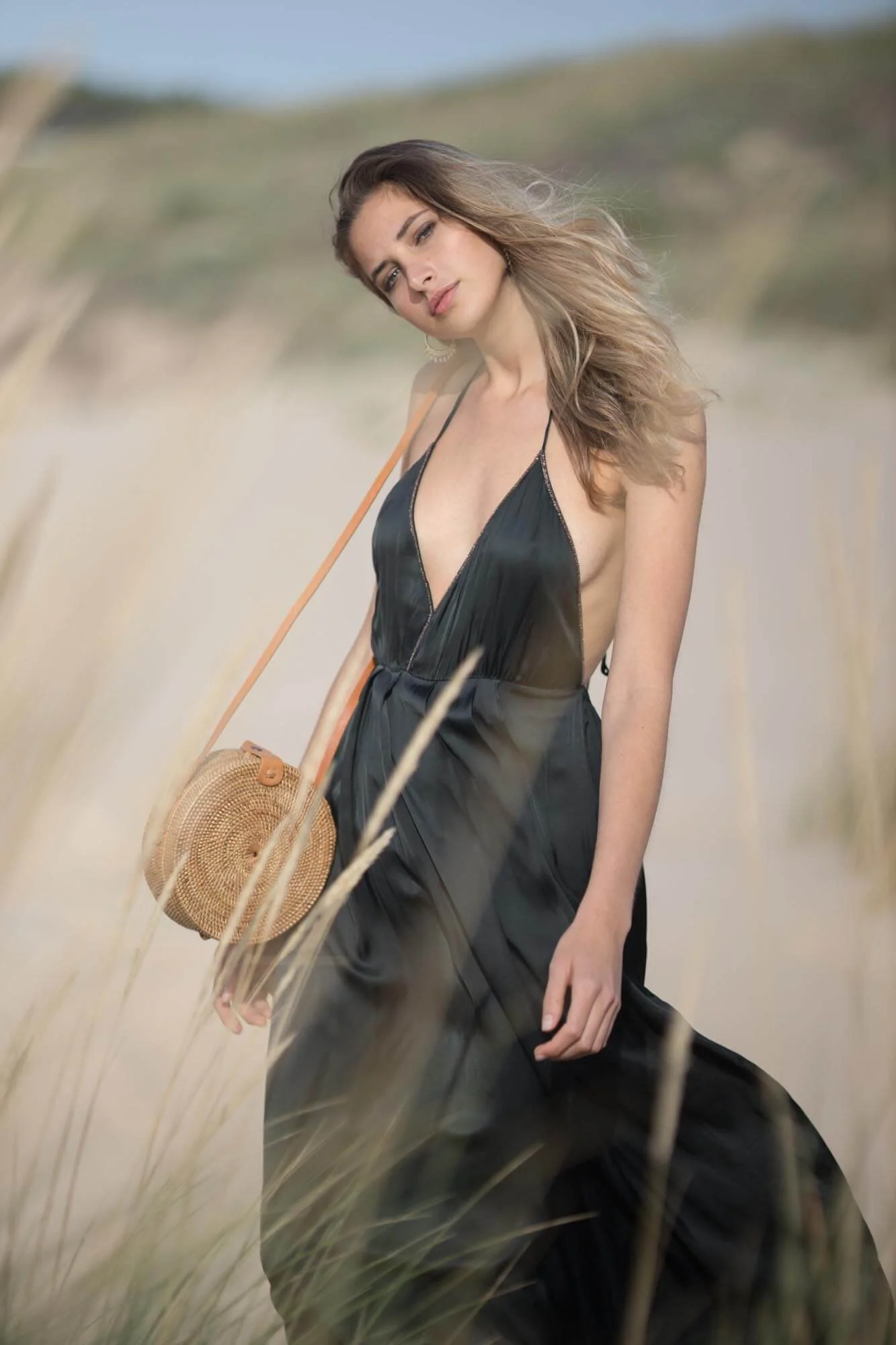 A woman in a black dress walking outdoors on a sandy path with tall grasses, holding a round rattan handbag.