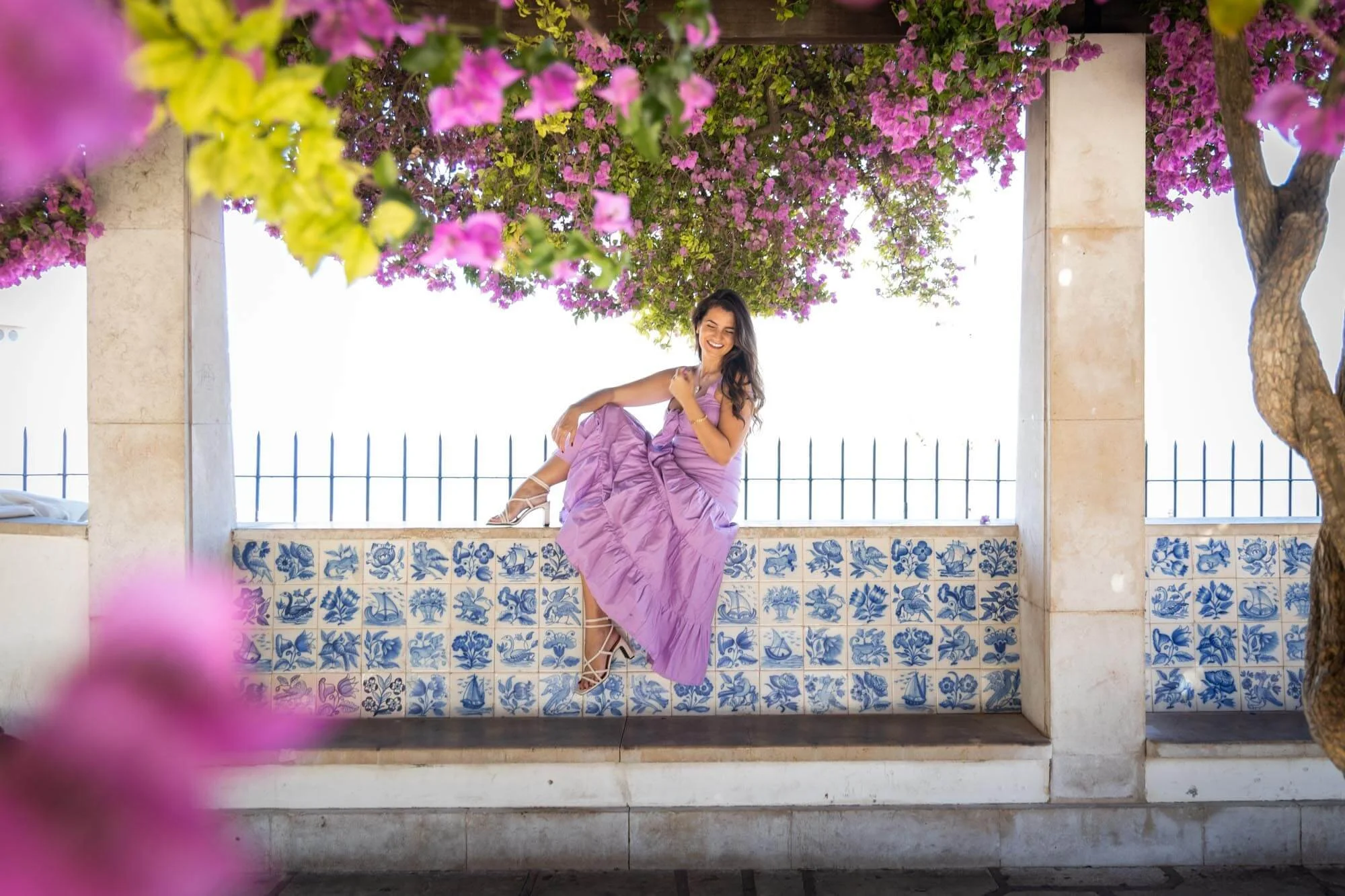 A woman in a purple dress and high heels sitting on a stone ledge under a flowering pink tree, smiling and enjoying the outdoors.