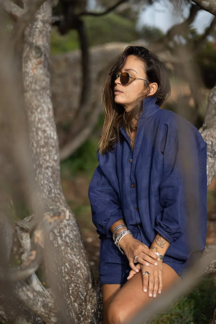 Woman wearing sunglasses and a blue jacket, sitting outdoors among trees with a contemplative expression.