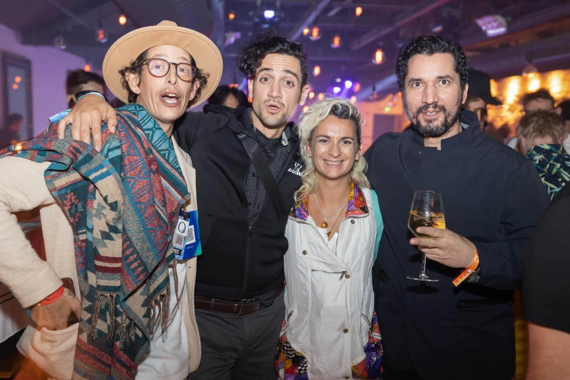 Group of four people smiling at a social event, with lighting and other attendees in the background.