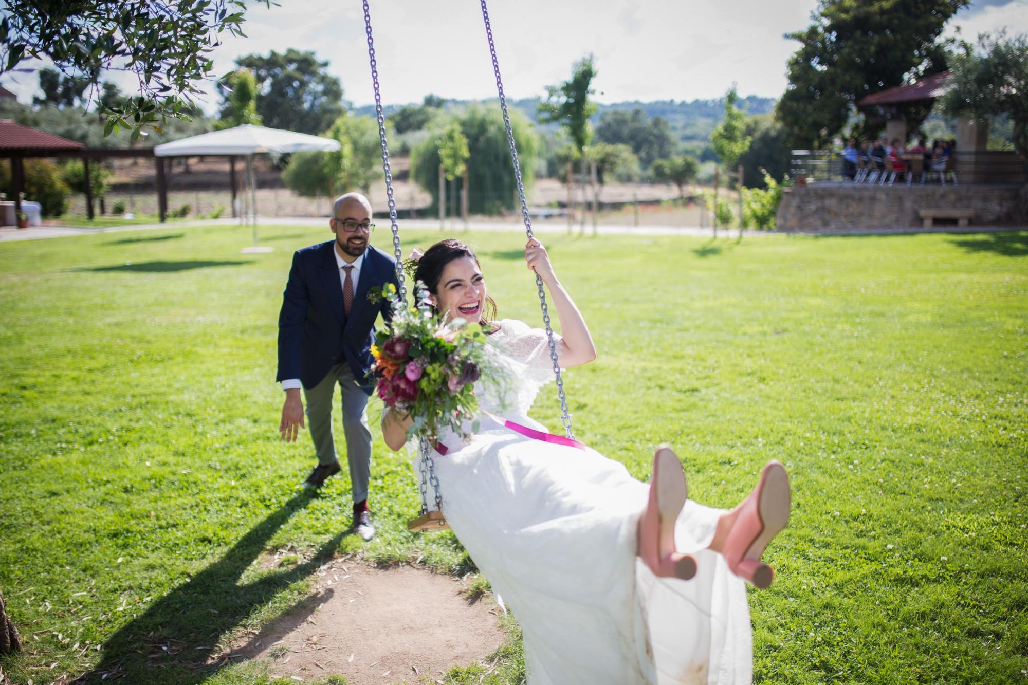 A bride in a white wedding dress sitting on a swing holding a bouquet of flowers, with a man in a blue suit pushing her, on a grassy lawn on a sunny day.
