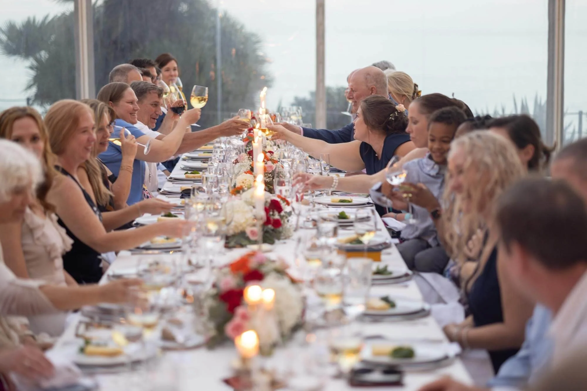 People at a wedding reception raising glasses in a toast, seated at a long decorated table with flowers and candles.