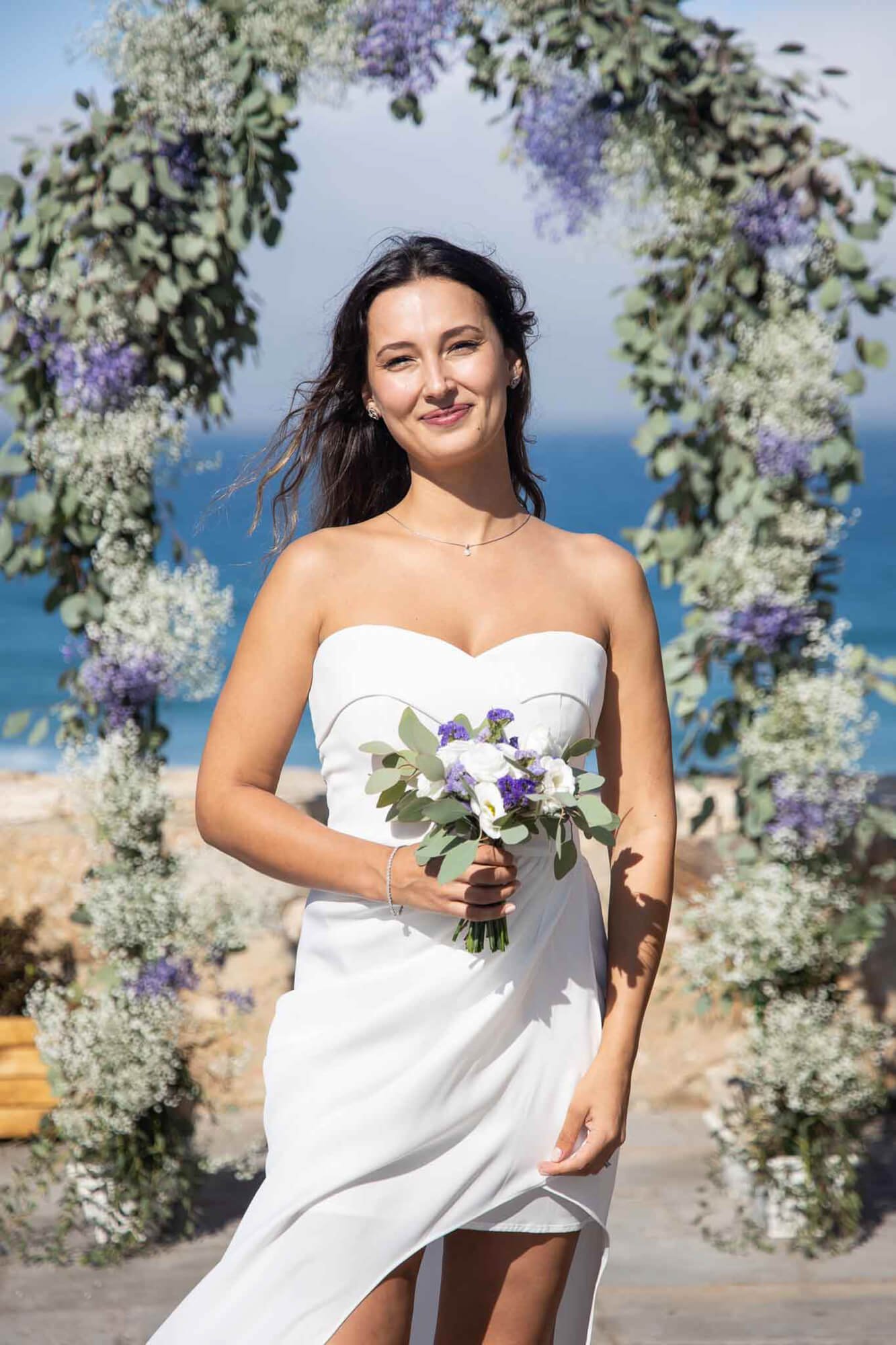 A bride in a white strapless wedding dress holding a bouquet of white and purple flowers standing in front of a floral wedding arch by the beach, with the ocean and partly cloudy sky in the background.