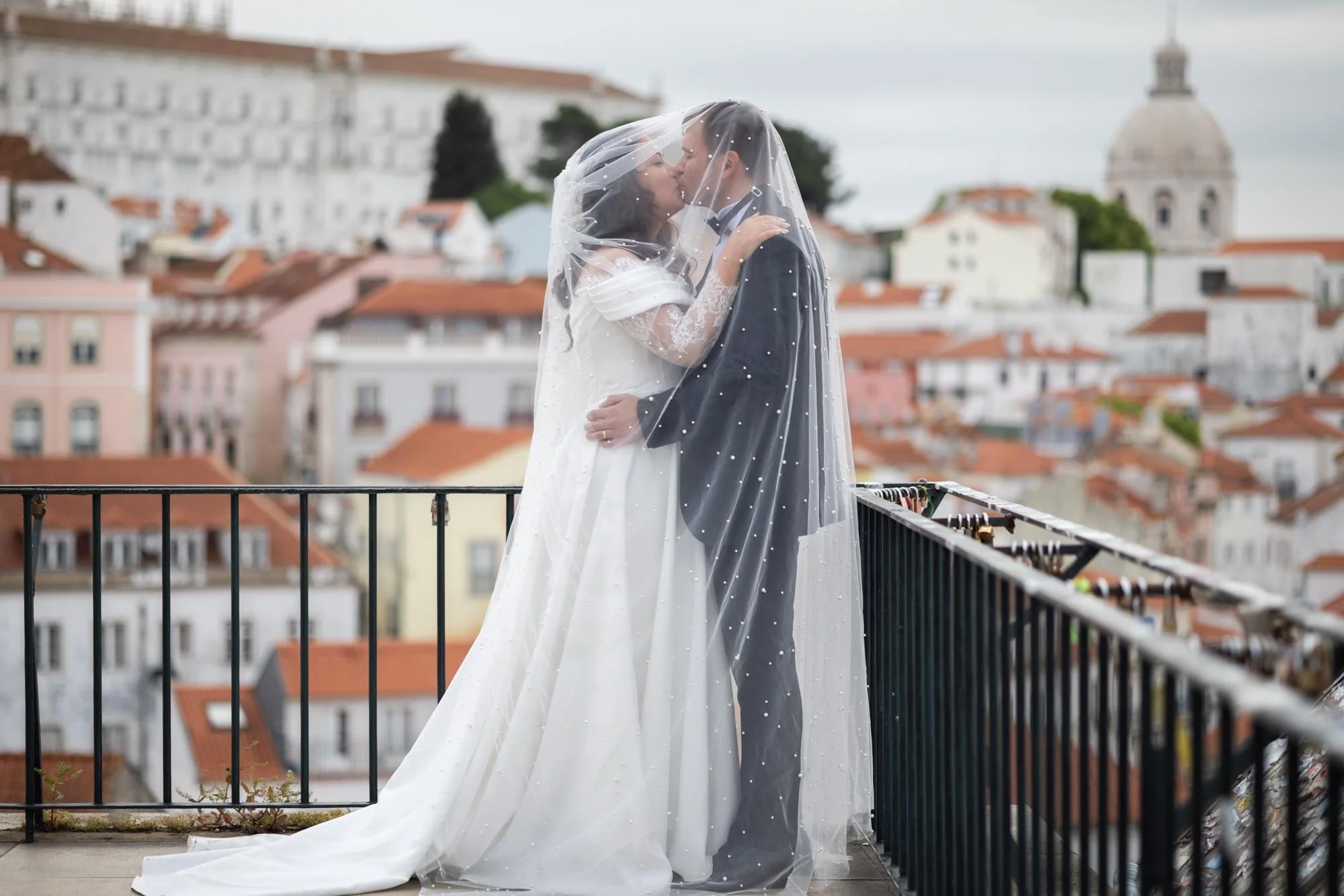 A newlywed couple kissing on a rooftop balcony with a cityscape and church dome in the background, both under a sheer wedding veil.