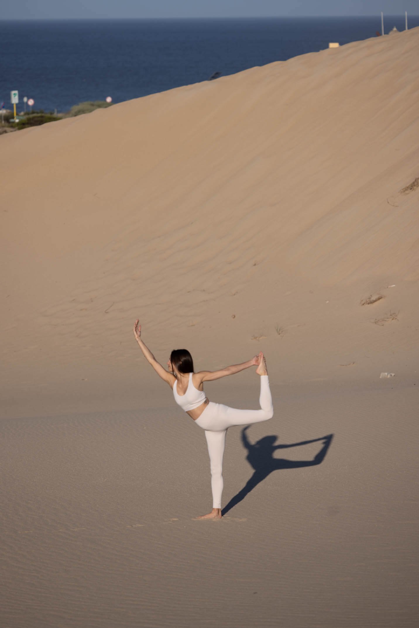 A woman practicing yoga in the desert, standing on one foot, holding her opposite foot behind her, with a large sand dune and the ocean in the background.