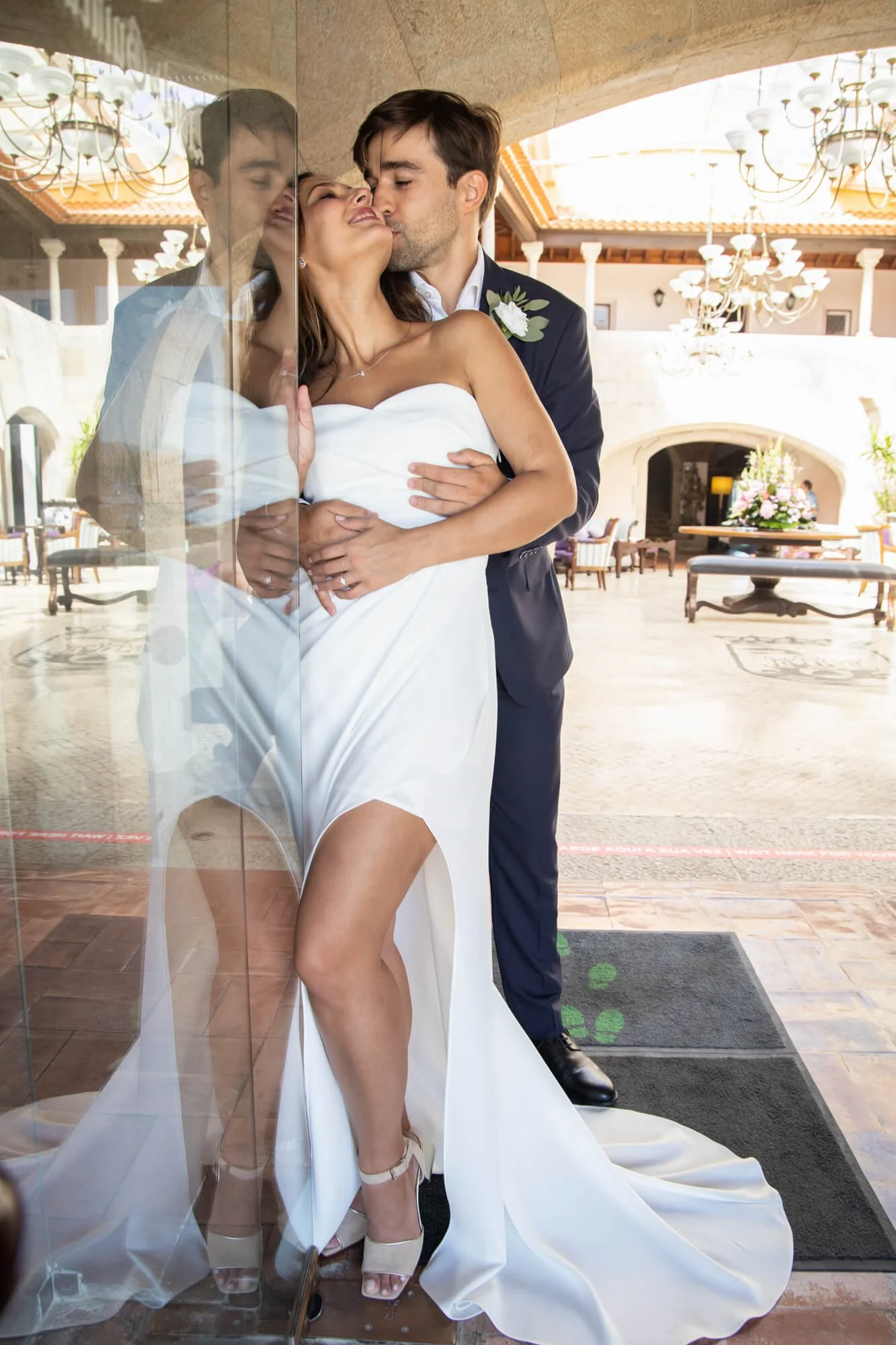 A bride and groom hugging in a hotel lobby, with the bride leaning against a glass wall, the groom kissing her cheek, and her wearing a white strapless wedding dress with high heels.