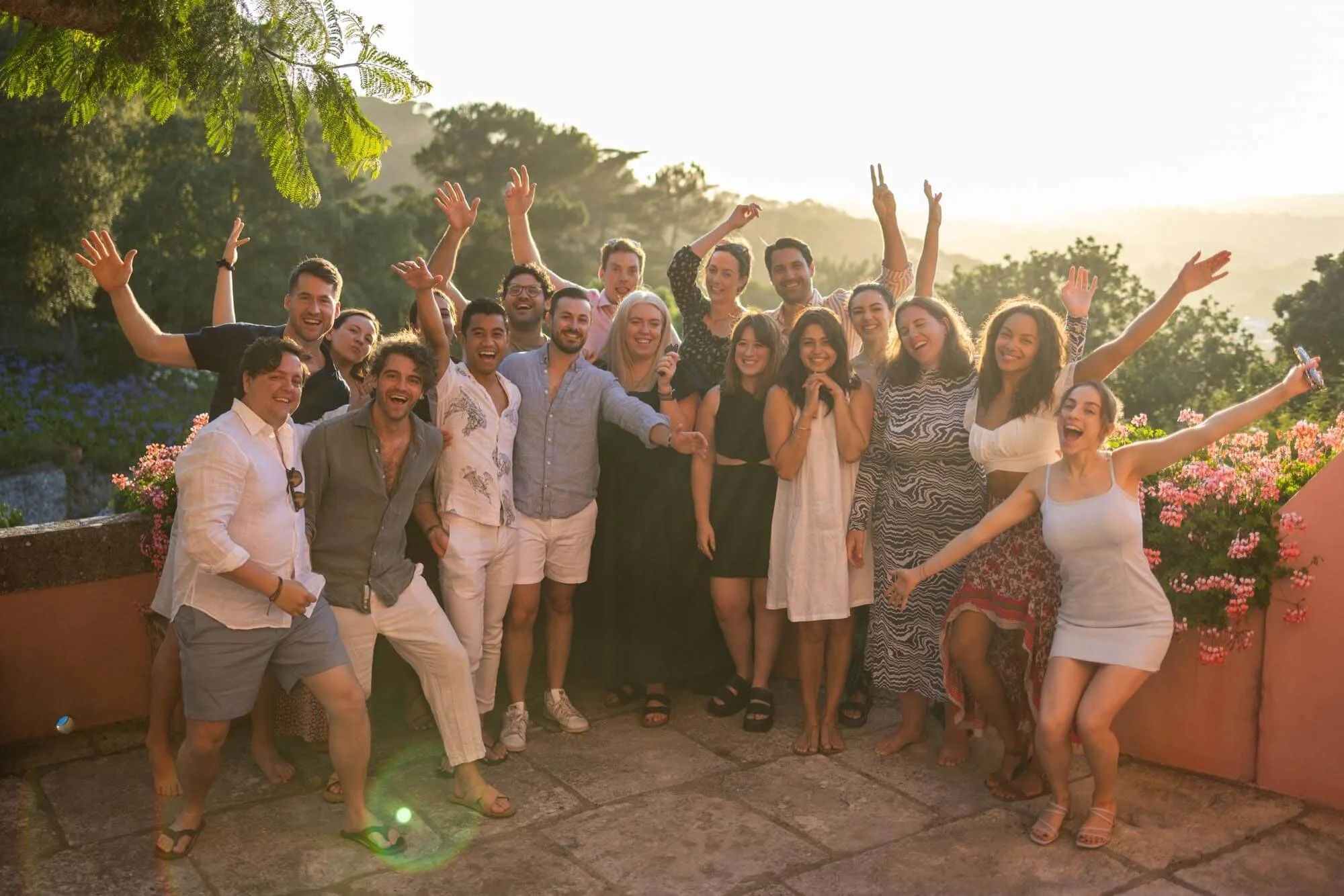 Group of people smiling and celebrating outdoors during sunset, standing on a stone patio with trees and flowers in the background.