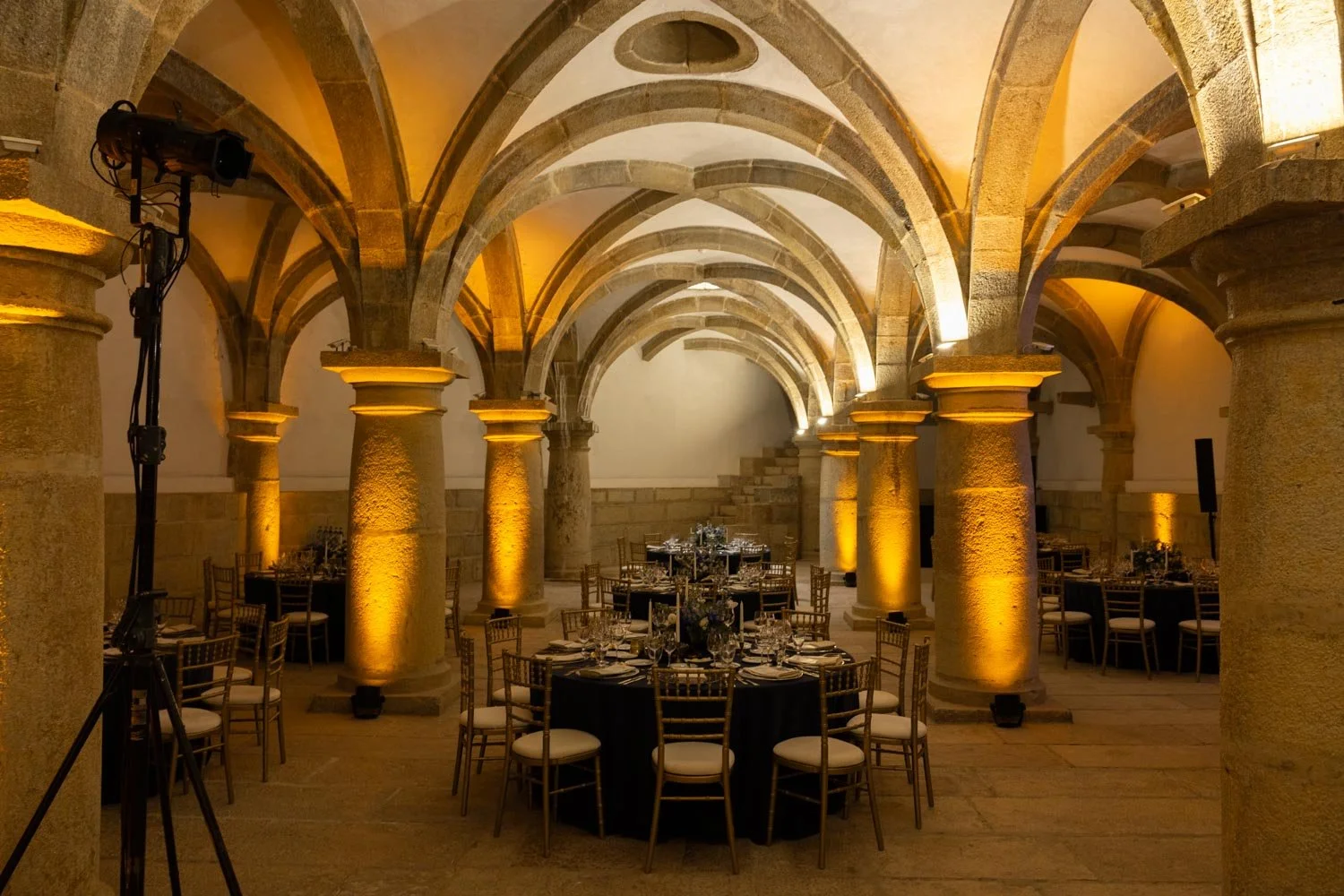 Elegant banquet hall with stone arches, columns, and warm yellow lighting, featuring round tables with black tablecloths and gold chairs set for an event.
