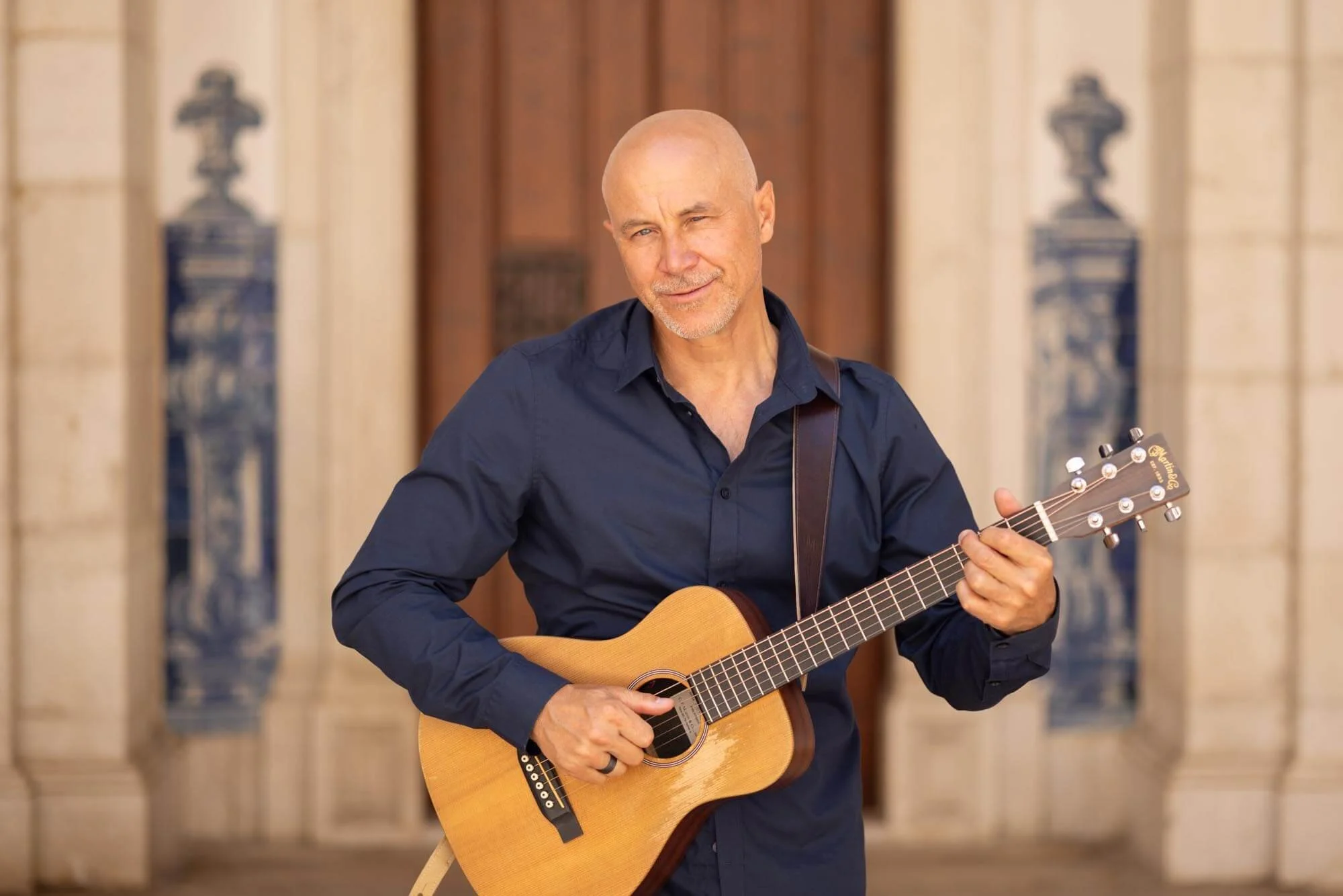 A bald man with a goatee in a dark blue shirt playing an acoustic guitar outdoors with historic architecture in the background.