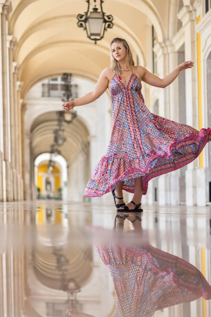 A young woman in a colorful, patterned sundress dances or twirls in an arched, open-air corridor with hanging lanterns, her reflection visible on a polished floor.