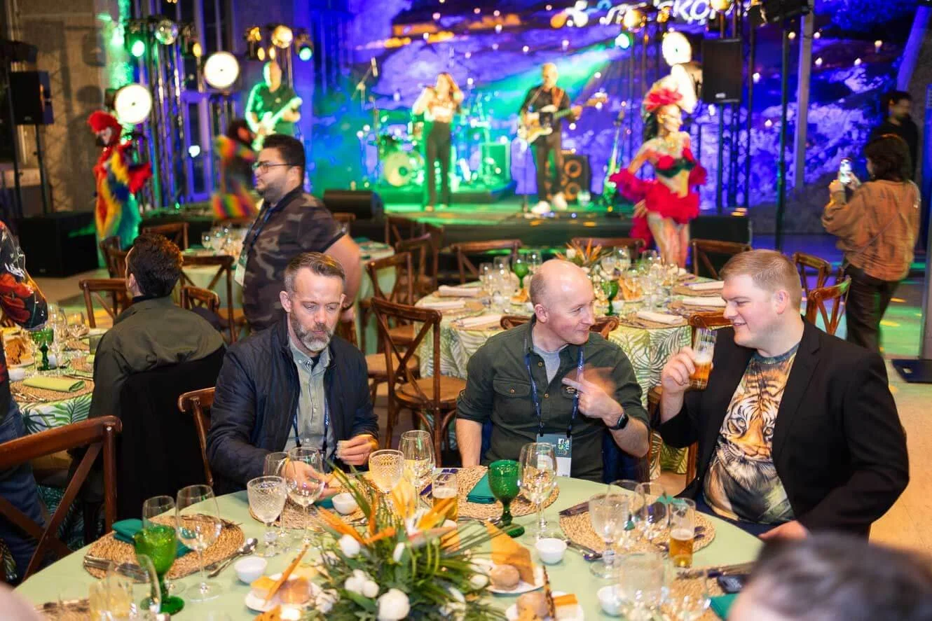 People sitting at a table with drinks and food in front of a stage where a band is performing. The band members are playing instruments, with some performers dressed in colorful costumes, on a decorated stage with colorful lights.