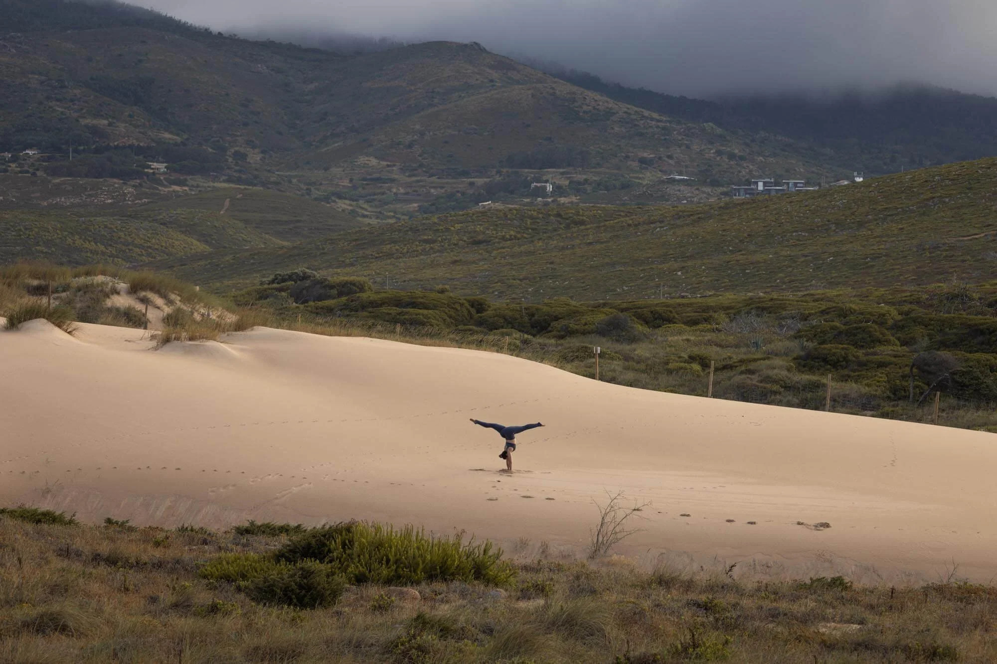 A person performing a handstand on a sandy dune in a desert landscape with green bushes, rolling hills, mountains, and cloudy sky in the background.