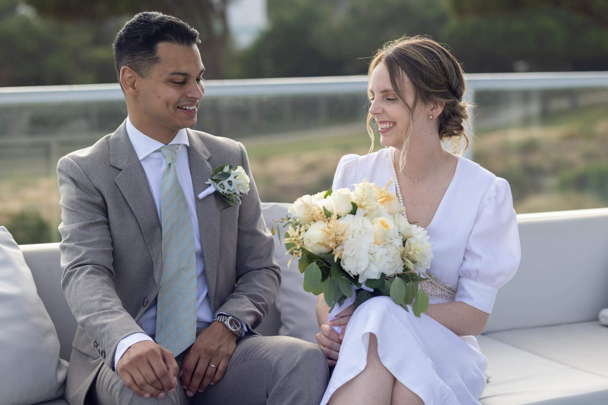 A bride and groom sitting on a white outdoor sofa, smiling at each other, with the bride holding a bouquet of white flowers, in a scenic outdoor setting.