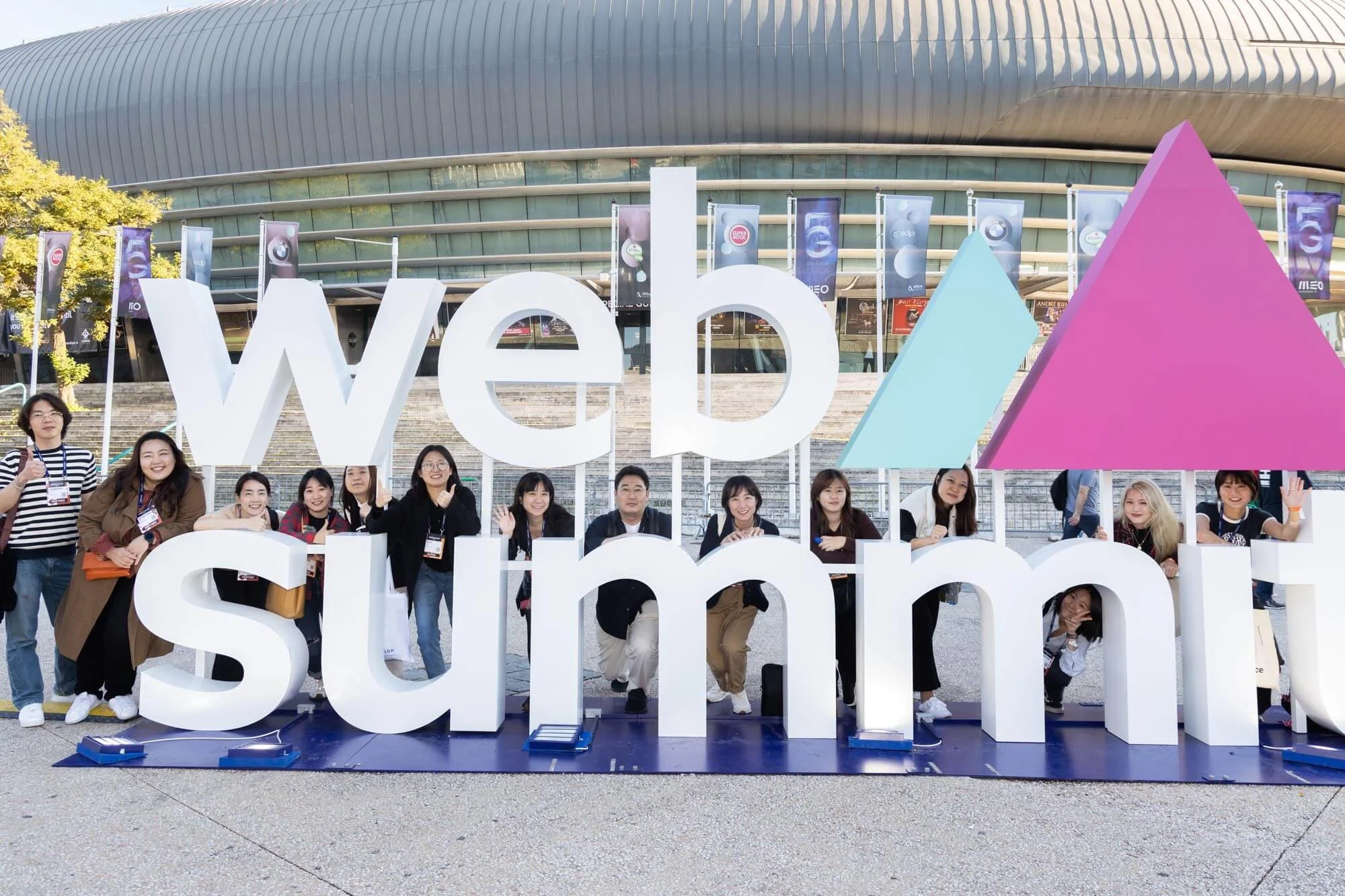 People posing behind large 3D sign that reads 'Web Summit' outside a modern building with banners.