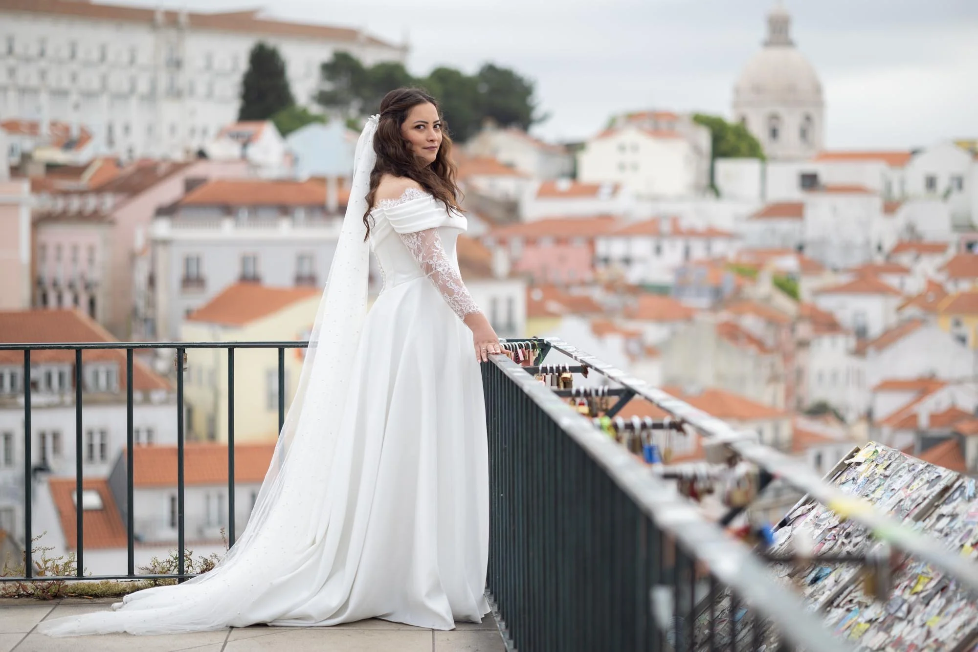 A woman in a white wedding dress with lace sleeves and an off-the-shoulder design, standing on a balcony overlooking a city with red-tiled roofs and historic buildings, with a domed structure in the background.