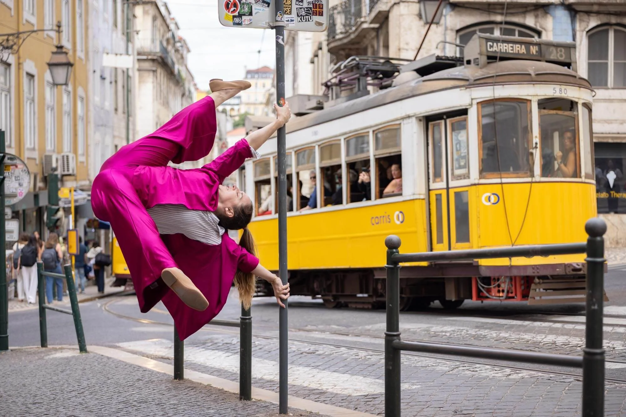A woman in a pink dress performing a pole dance move on a street barrier in front of a yellow tram, with onlookers inside the tram and pedestrians on the sidewalk.