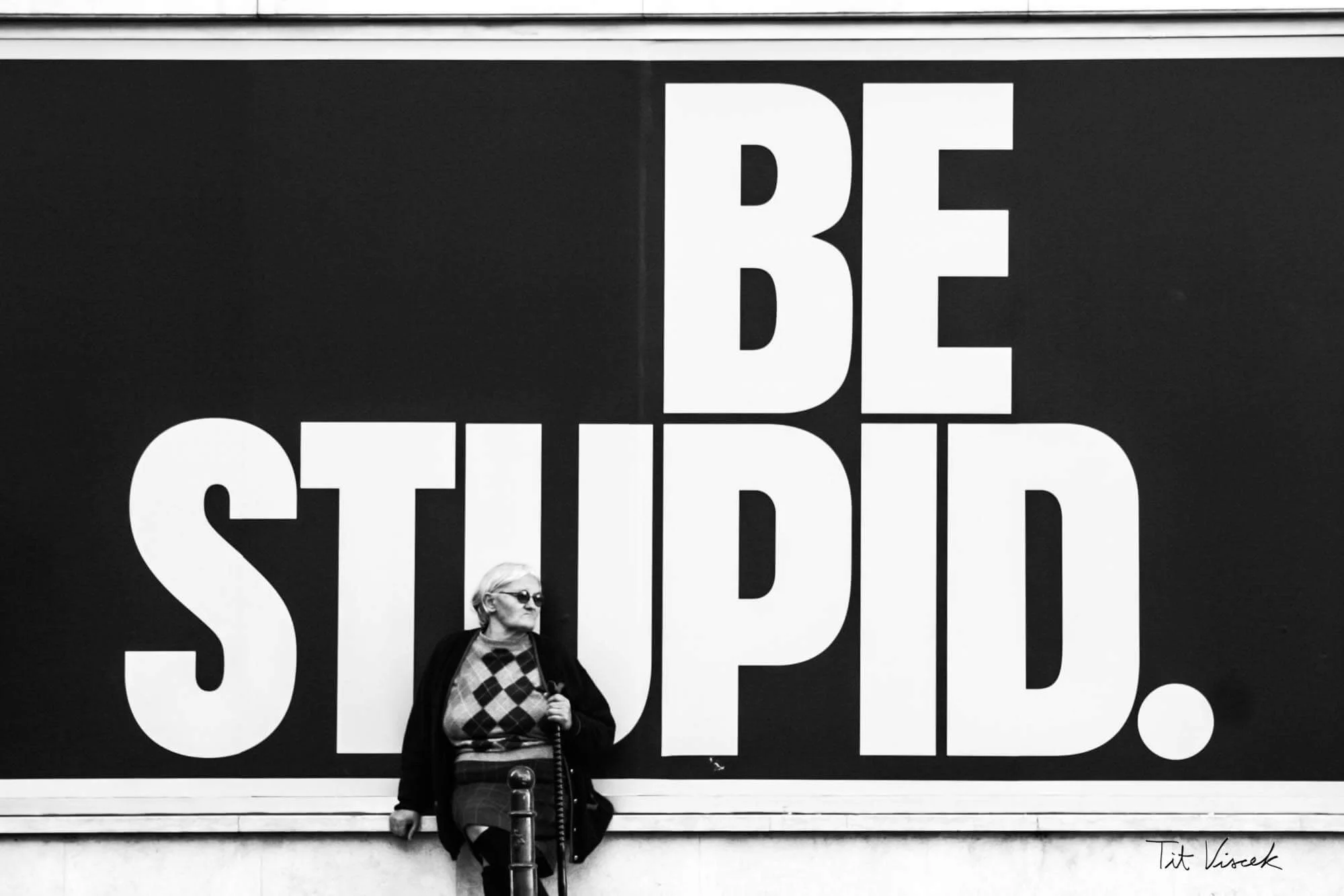 A woman wearing glasses, a patterned sweater, and a coat stands in front of a large black billboard with the white text "BE STUPID."
