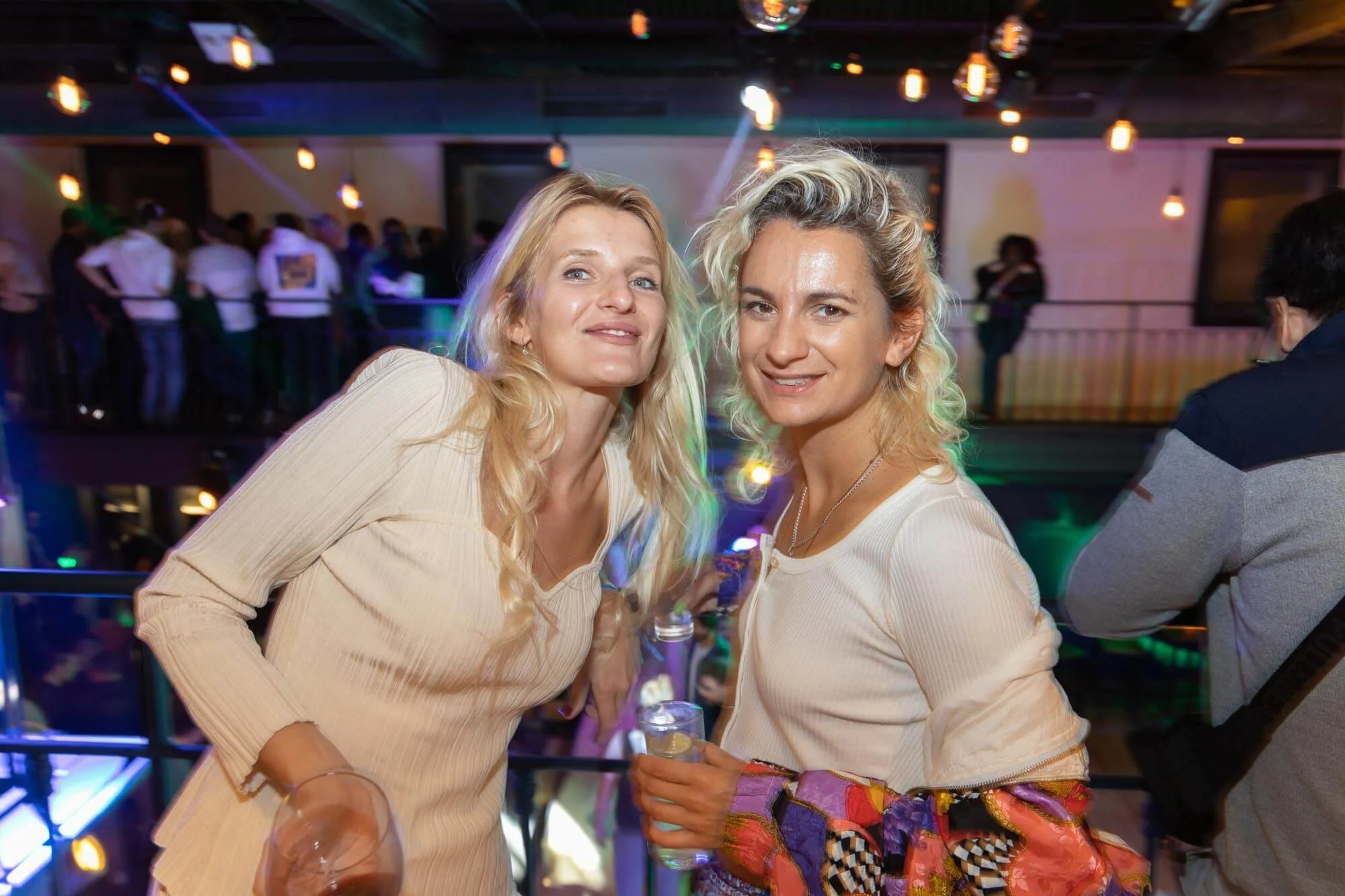Two women with blonde hair at a party or nightclub, smiling at the camera, one holding a drink. Background shows other people, colorful lights, and a lively atmosphere.