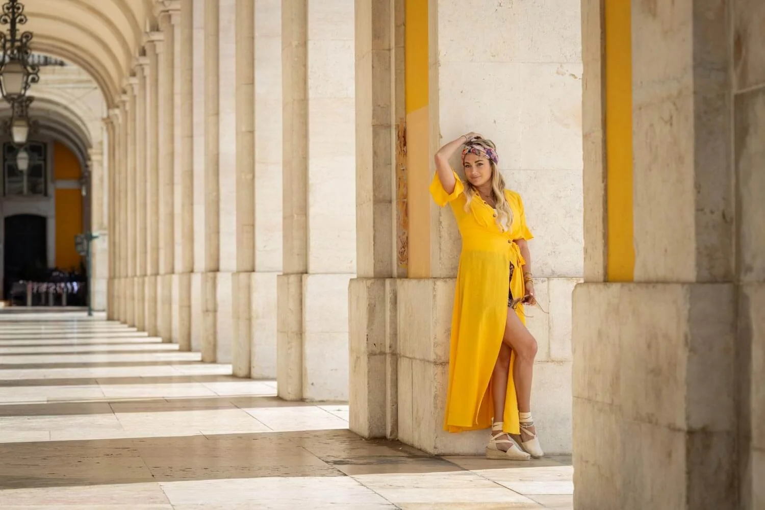 A woman in a yellow dress and beige shoes standing in a colonnade with stone arches and columns, touching her head with one hand and holding sunglasses in the other.