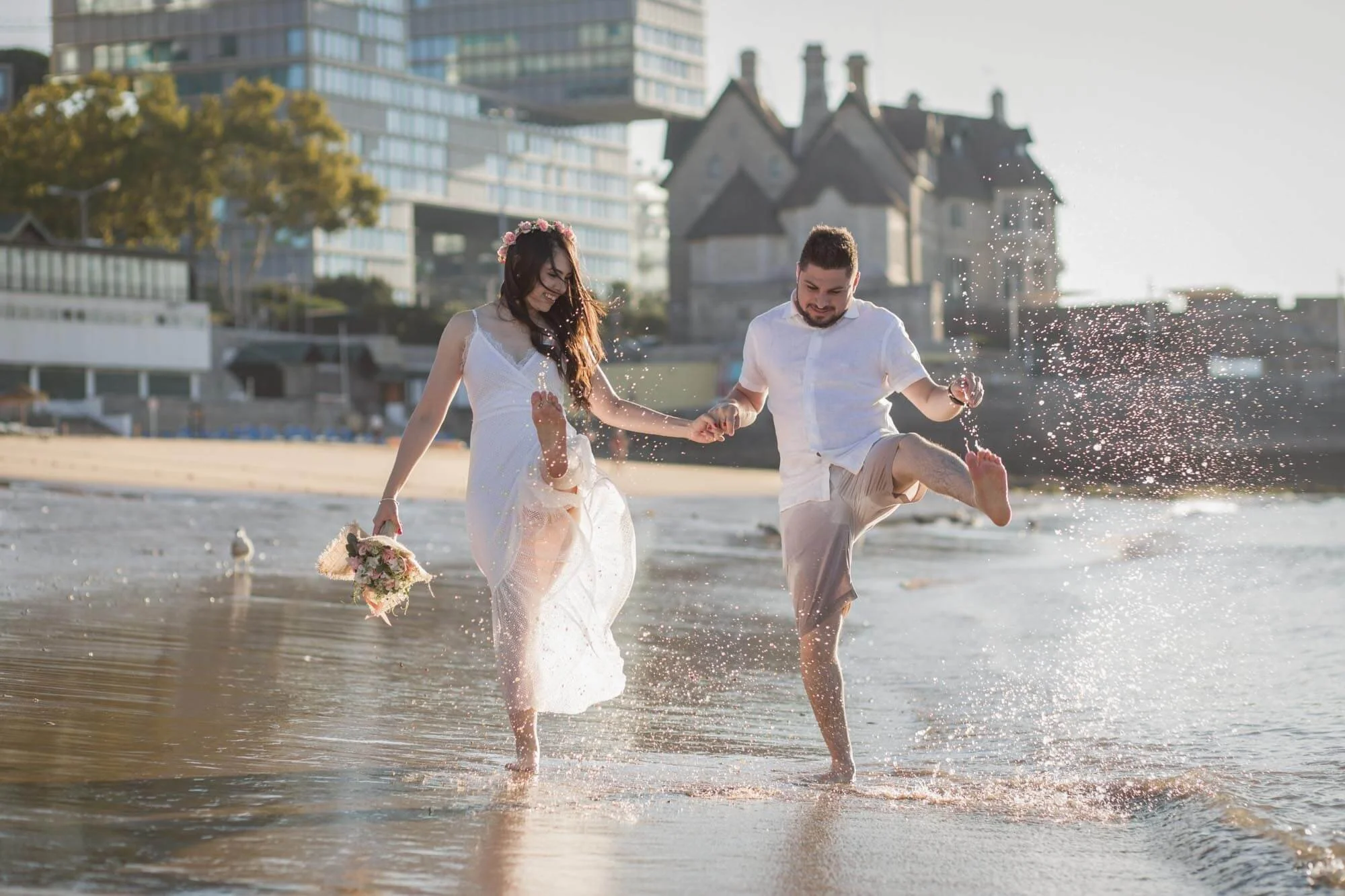 A couple dressed in white clothes holding hands and kicking water on the beach during sunset, with buildings and trees in the background.