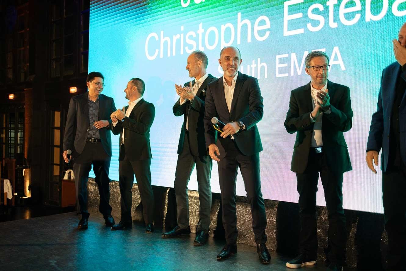 Six men in suits standing on a stage in front of a large screen that displays the name 'Christophe Este' and 'South EM'. The men are smiling, clapping, or holding microphones.