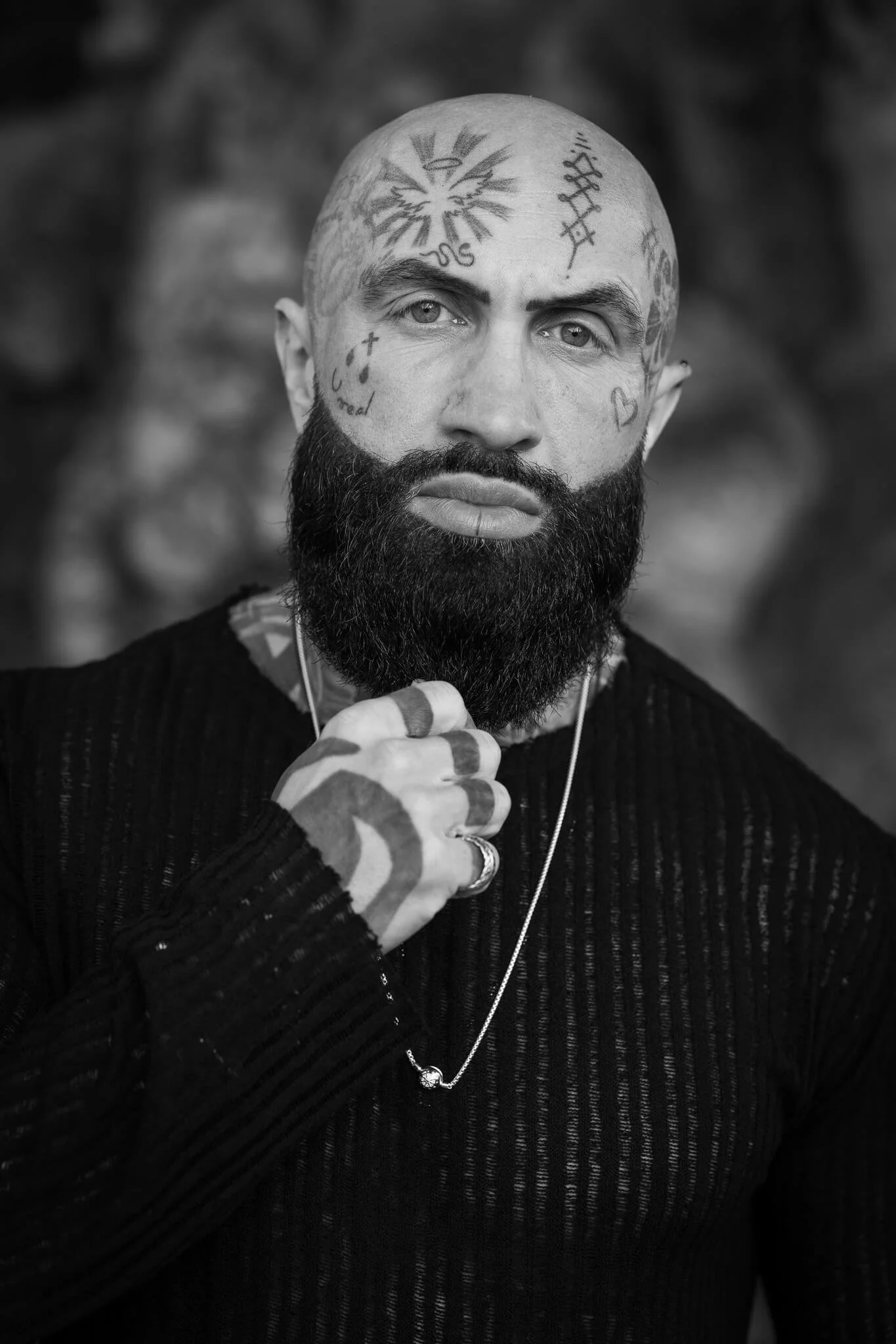 A black and white portrait of a man with a thick beard and numerous tattoos on his face, head, and hand. He is wearing a black, textured long-sleeve shirt and has a necklace around his neck. He is resting his chin on his hand, looking directly at the