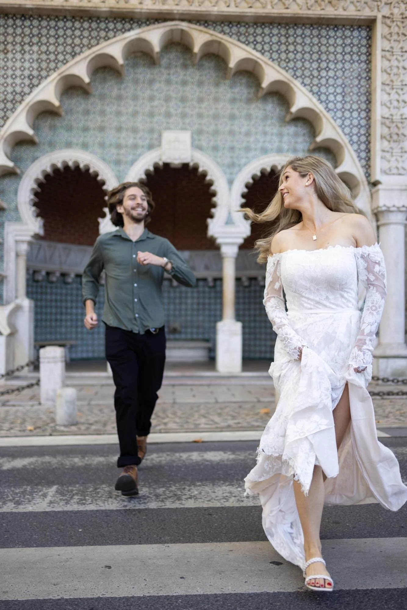 A woman in a white wedding dress walking across a crosswalk while smiling, followed by a man in a gray shirt and black pants who is running and smiling, in front of an ornate, colorful building with arches.