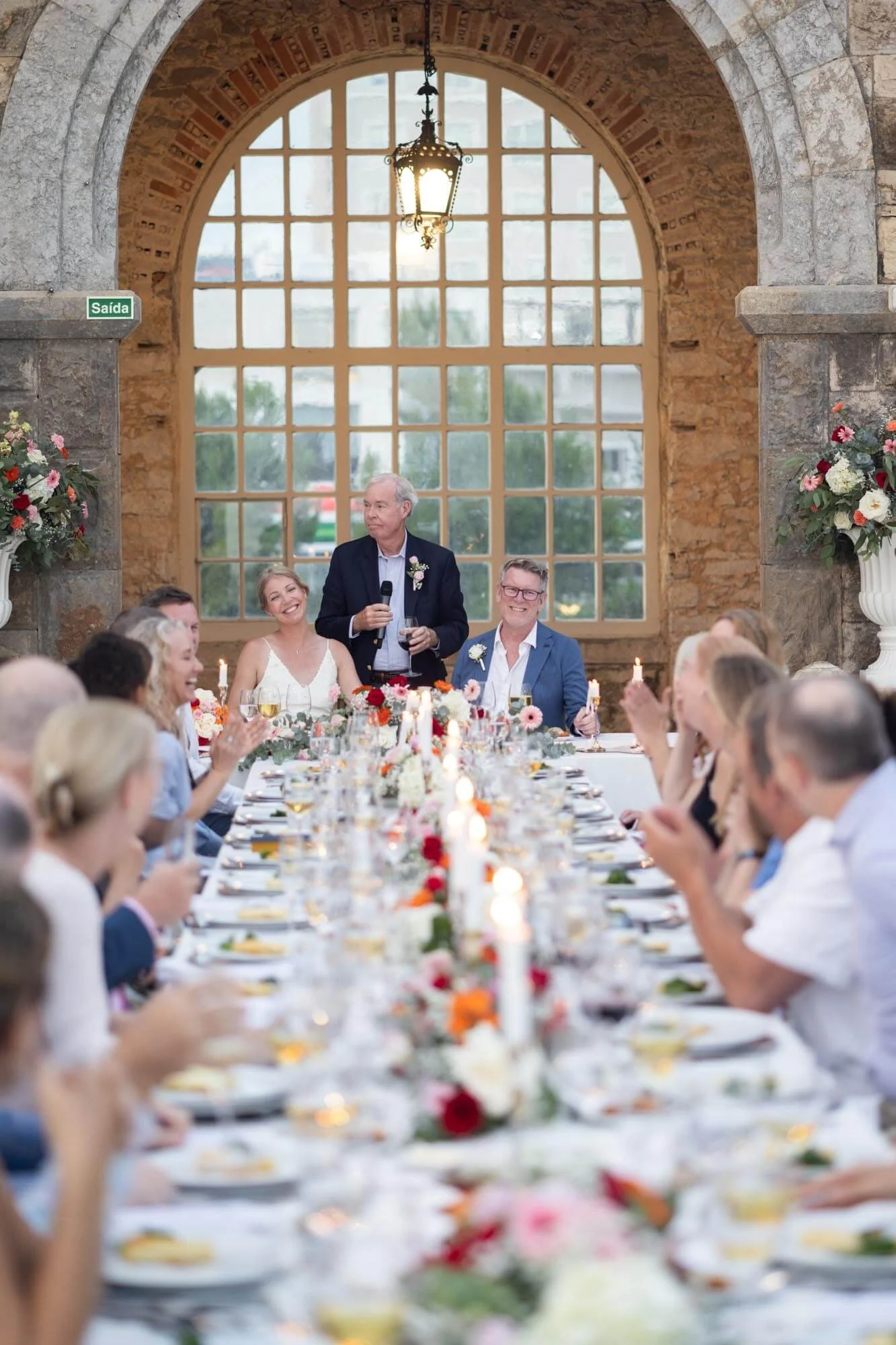 A wedding reception with a long table decorated with flowers and candles, where guests are seated and listening to a toast. A man is standing with a microphone, speaking, with a woman in a white dress and man in a blue suit smiling nearby. The backgr