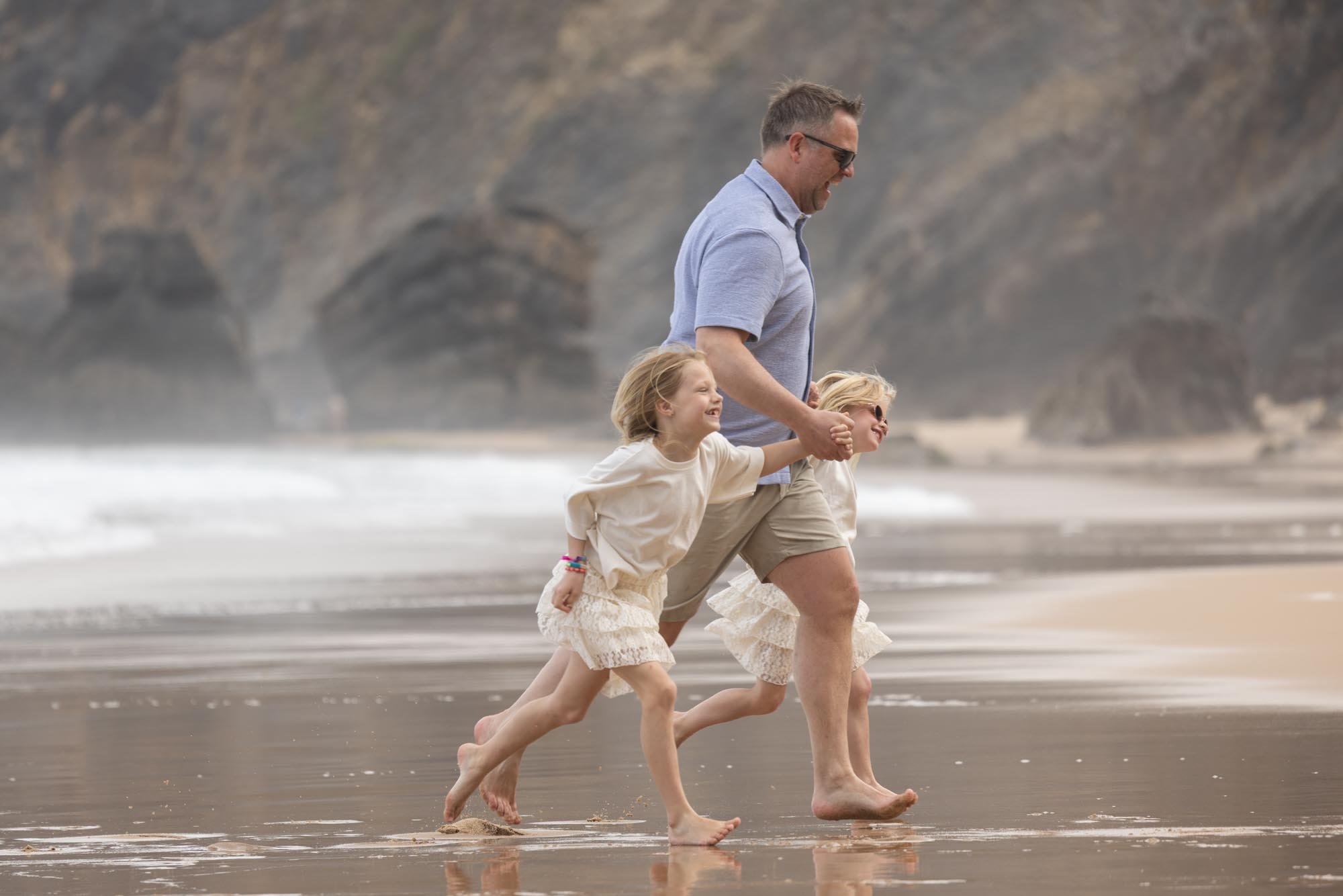 A man walking hand-in-hand with two young girls on a beach, smiling and enjoying a day near the ocean with cliffs in the background.