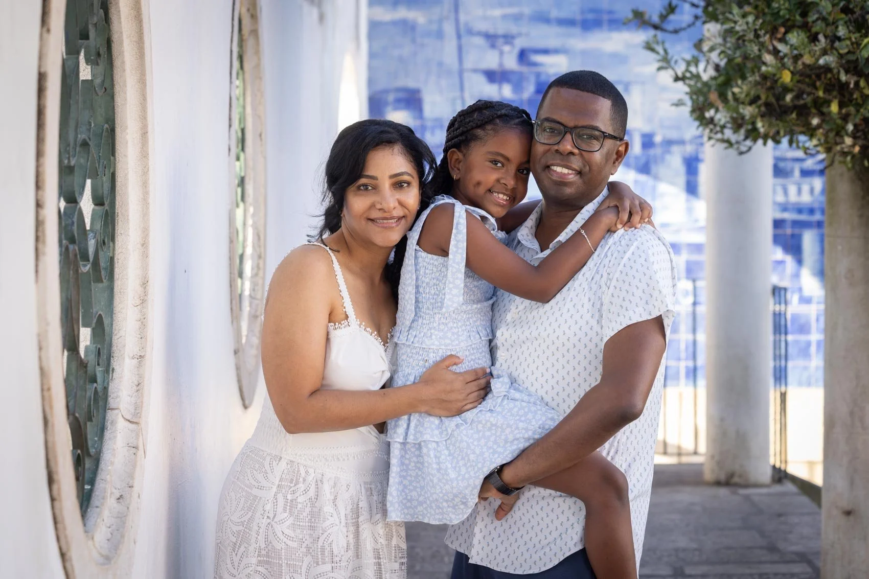 A family of three standing outdoors, smiling and embracing each other. The woman is on the left, wearing a white dress, the man is on the right, holding a young girl in his arms. The girl is smiling and wearing a light blue dress. The background show