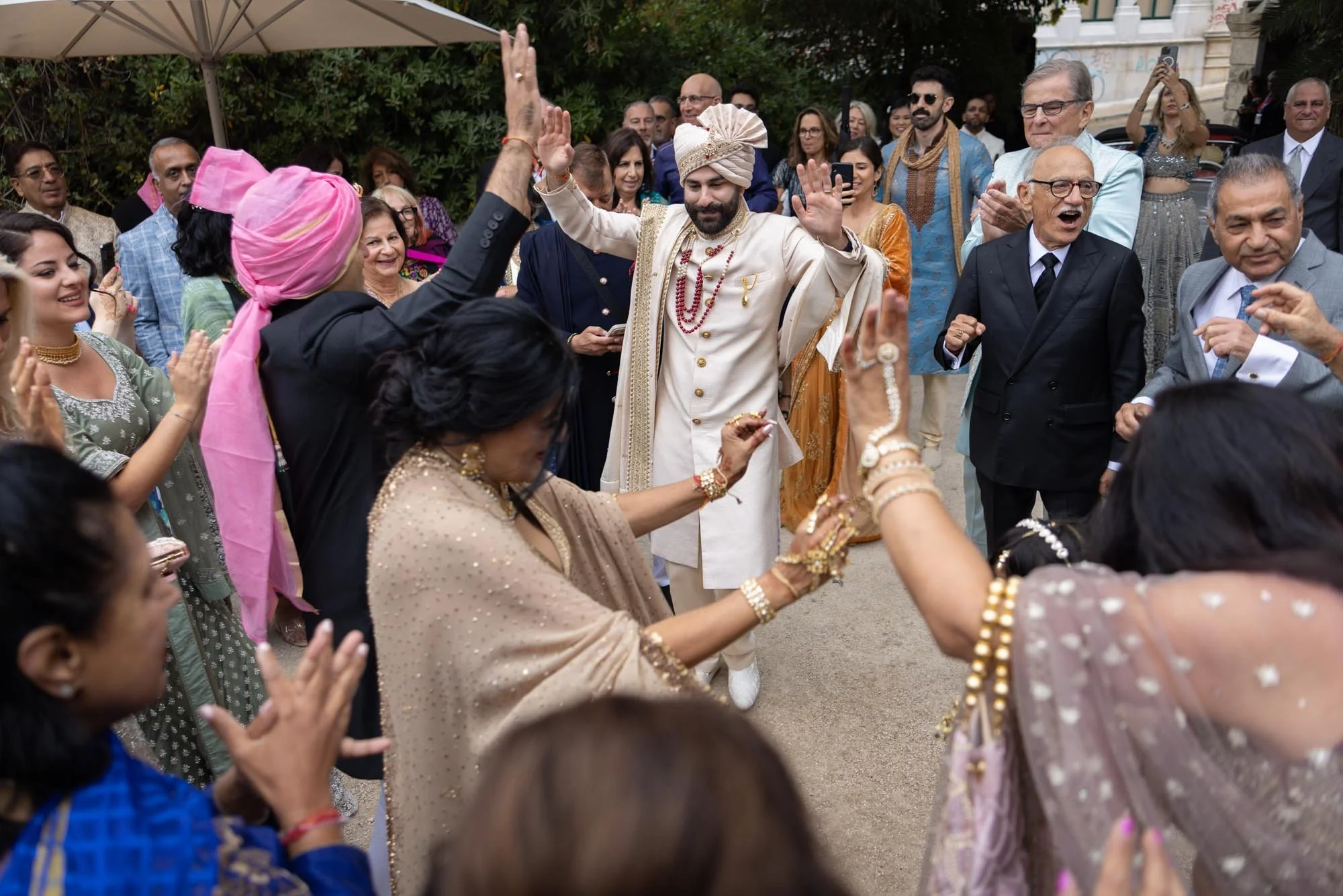 A group of people celebrating at a wedding, with men and women dressed in traditional Indian attire. The groom, in cream-colored attire with a turban, is dancing and smiling while surrounded by guests dancing and cheering.