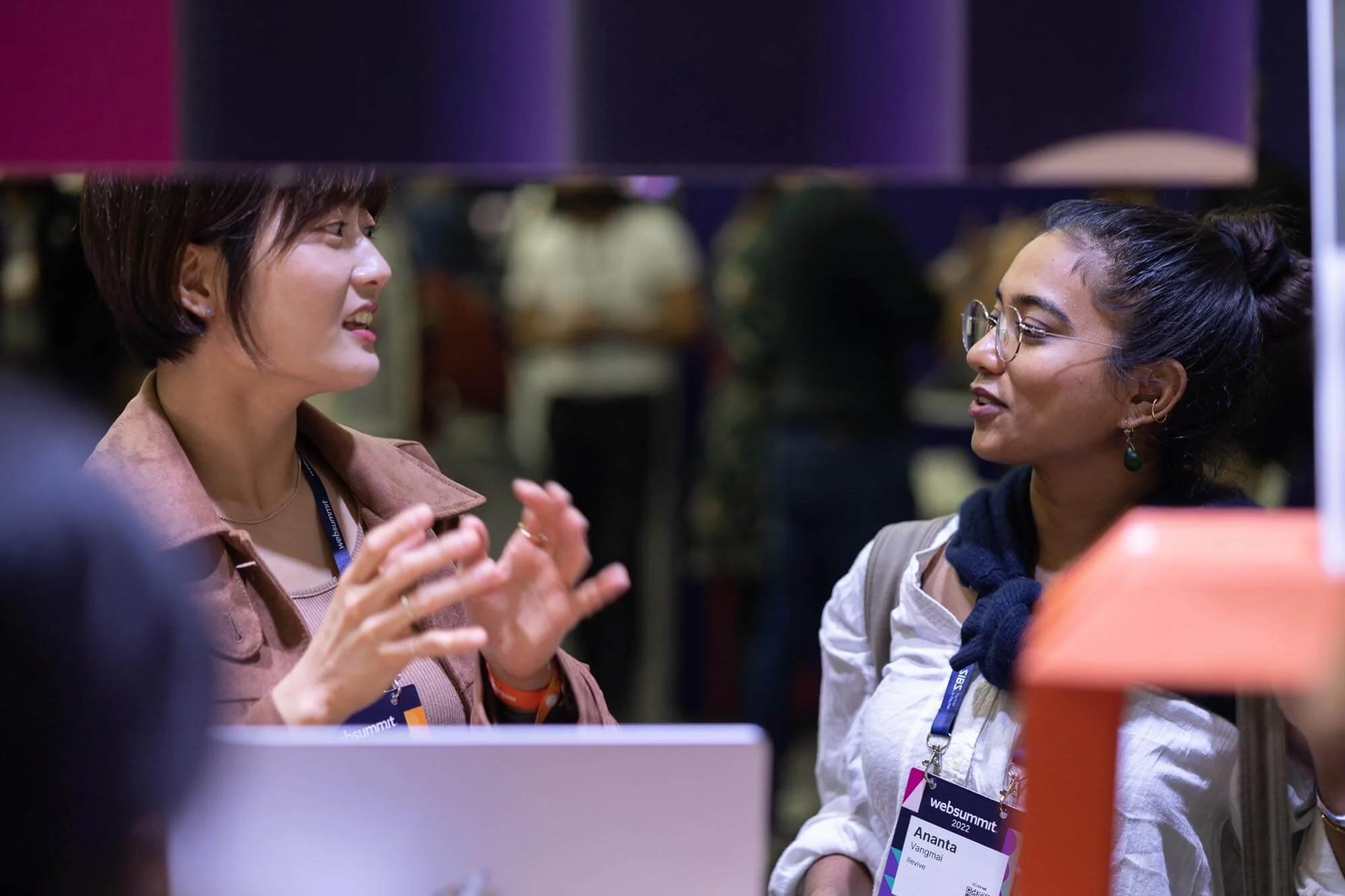 Two women engaged in conversation at a professional event, wearing name tags with 'websummit 2022' visible, in an indoor setting with other people in the background.