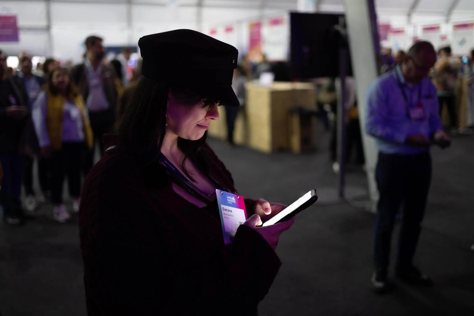 A woman in a black hat and maroon sweater looking at her phone in a crowded indoor event space, some people are in the background.