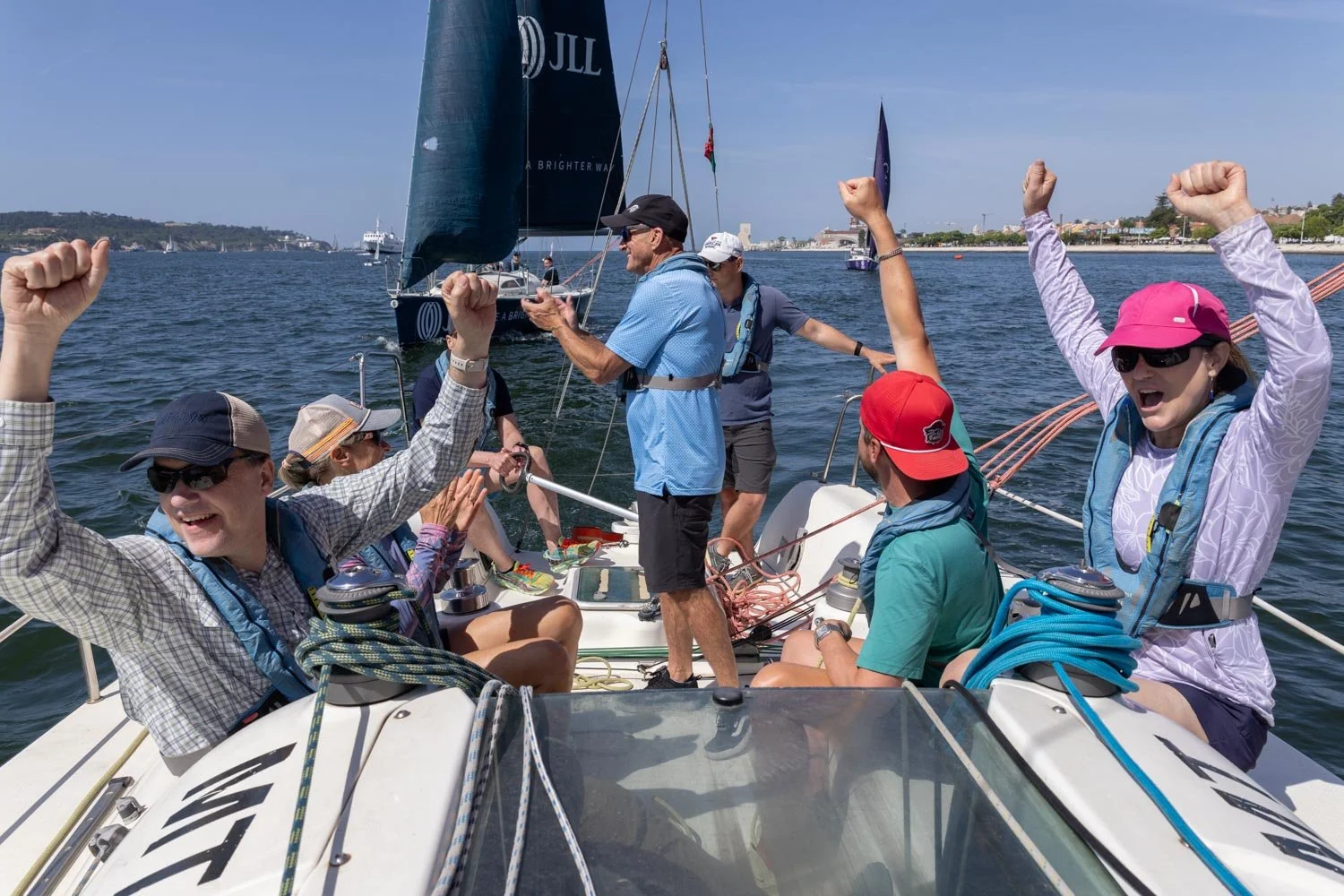 A group of people celebrating a win on a sailboat, raising their fists, with a man in a blue shirt taking photos. The scene is on the water with clear skies and other boats in the background. It is a corporate event , covered by Lisbon Event Photogra