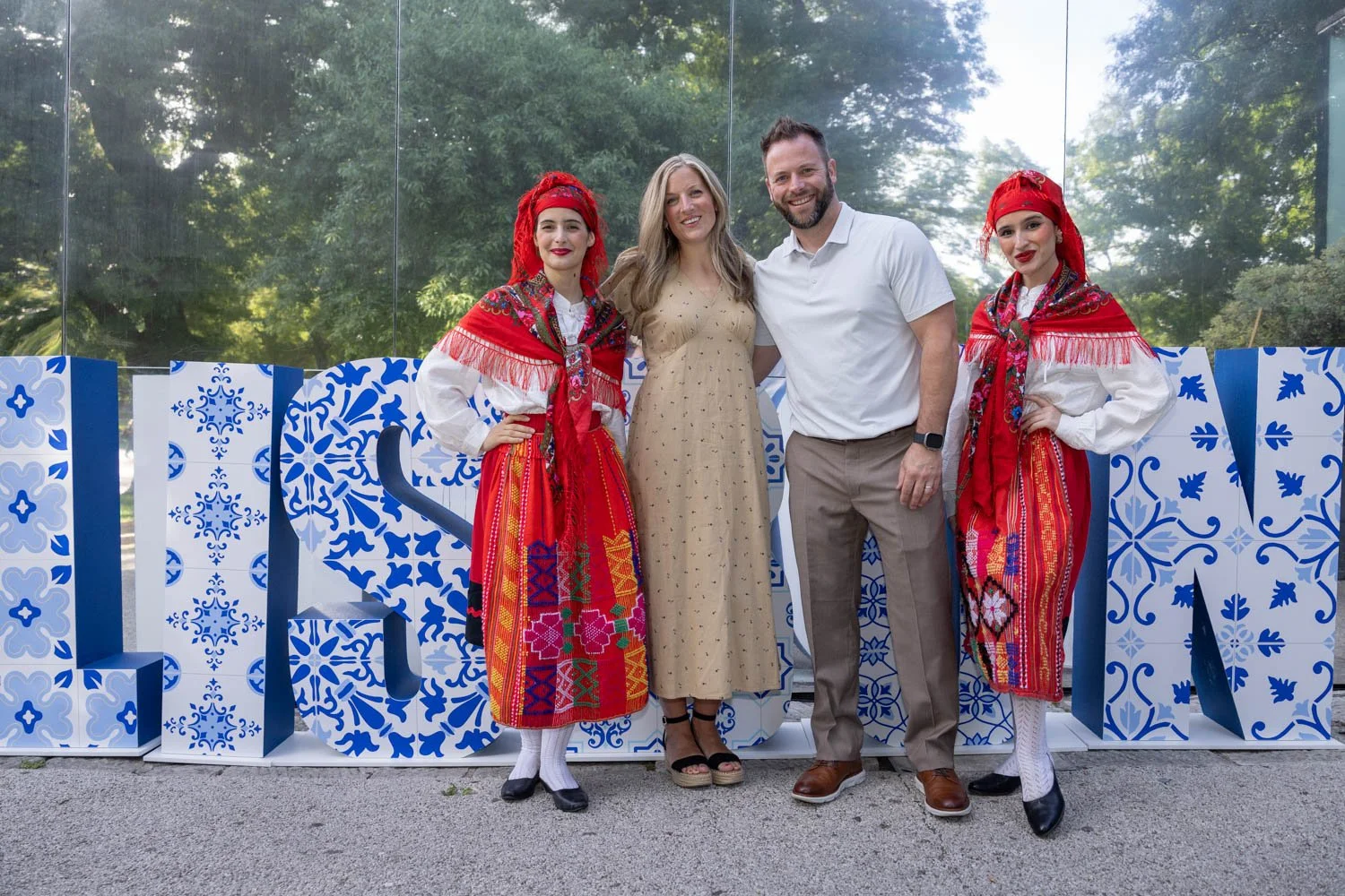Four people standing together in front of a decorative backdrop featuring blue and white patterns. The two women on the left and right are dressed in traditional Portuguese folk costumes with red headscarves, colorful skirts, and white blouses. The w