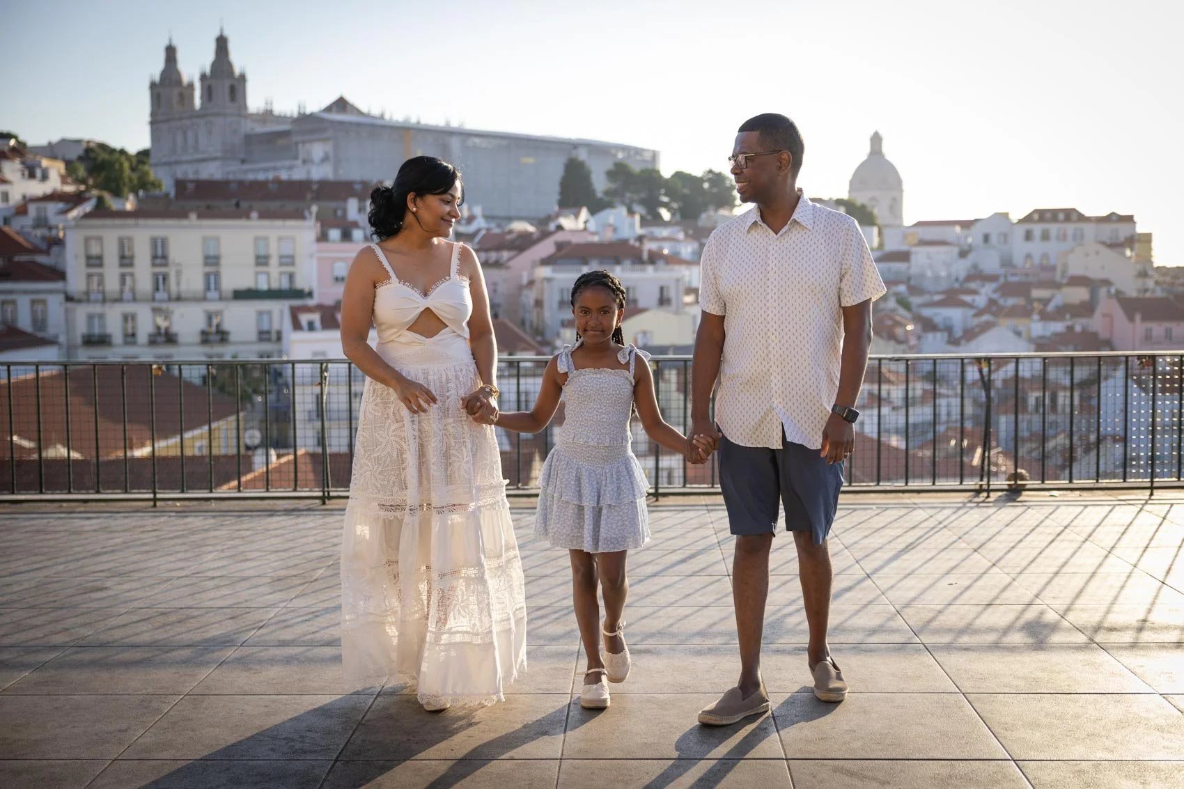 Family of four holding hands and walking at Miradouro Portas de Sol  with cityscape in the background at sunset.