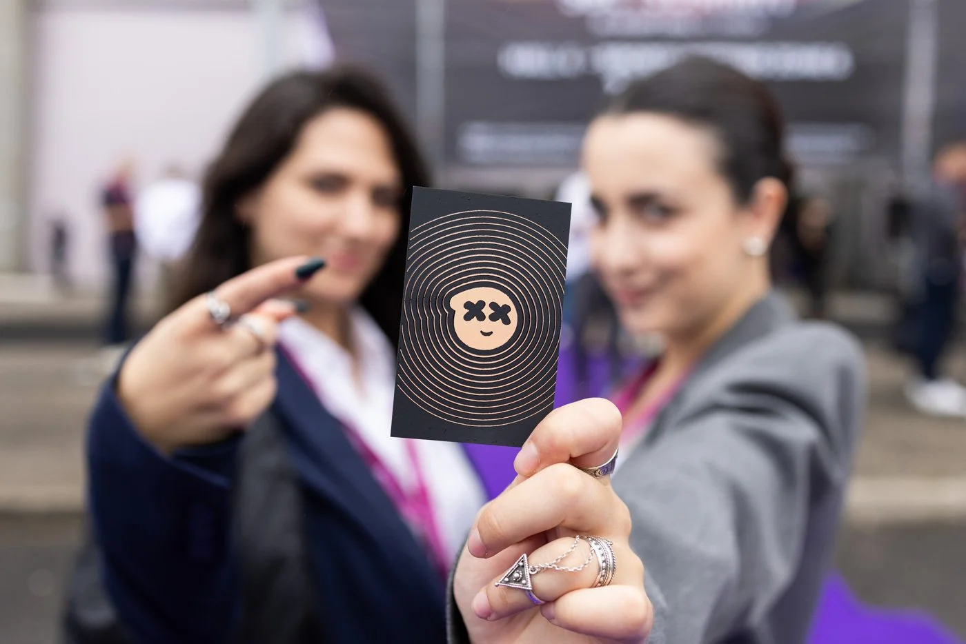 Two women in business attire holding up a black card with a stylized smiling face with X's for eyes and concentric circles around it, at a busy event or conference.